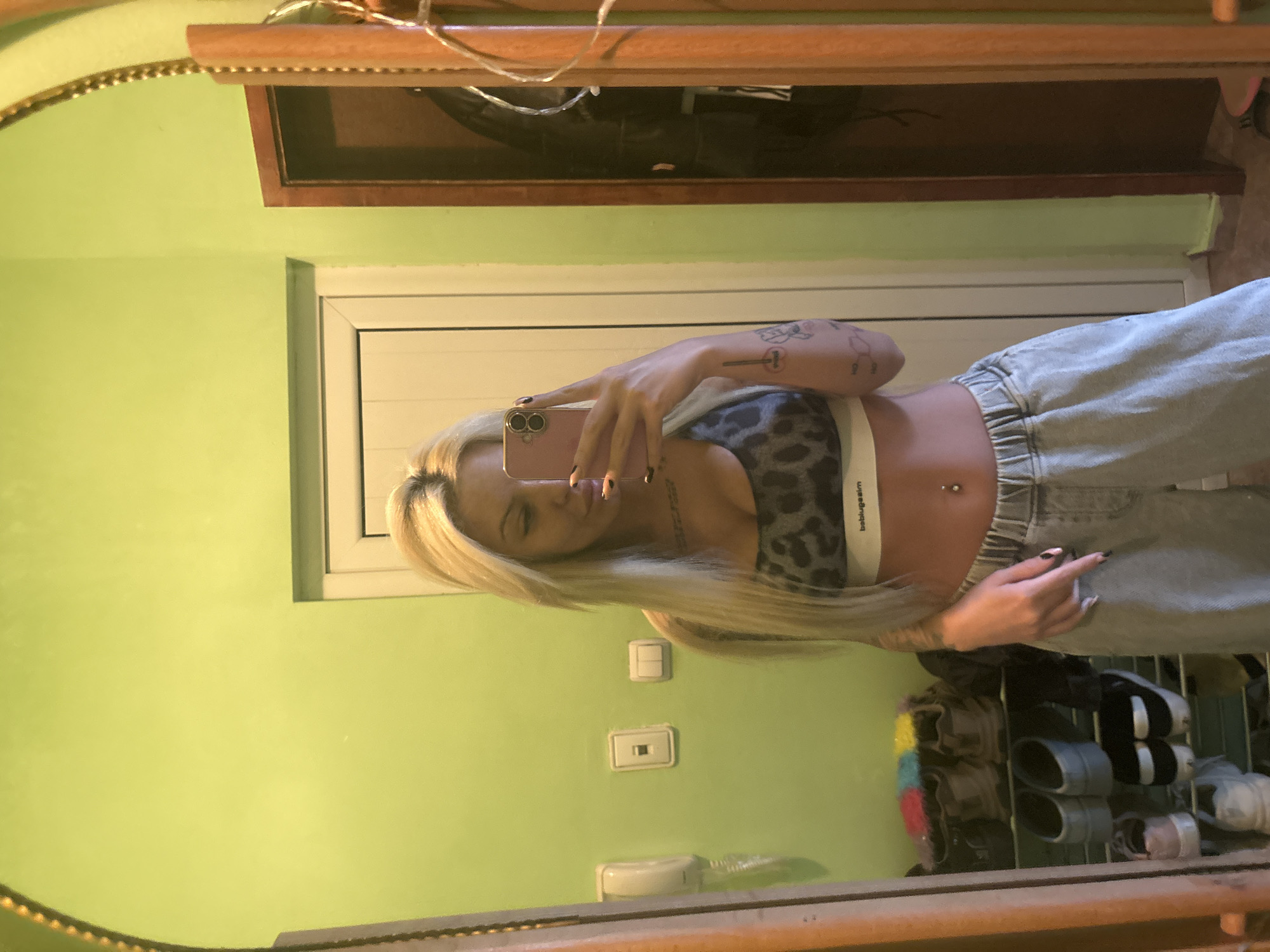 A woman is standing in an Unknown, Unknown location, taking a selfie in a mirror. The background wall is painted green and features a white light switch and a white wall socket. The woman has blonde hair and is wearing a gray leopard-print sports bra with a white elastic band. She is also wearing light-wash denim jeans. A mirror frame is visible in the foreground. Below the woman, a shoe rack containing various pairs of shoes is present.