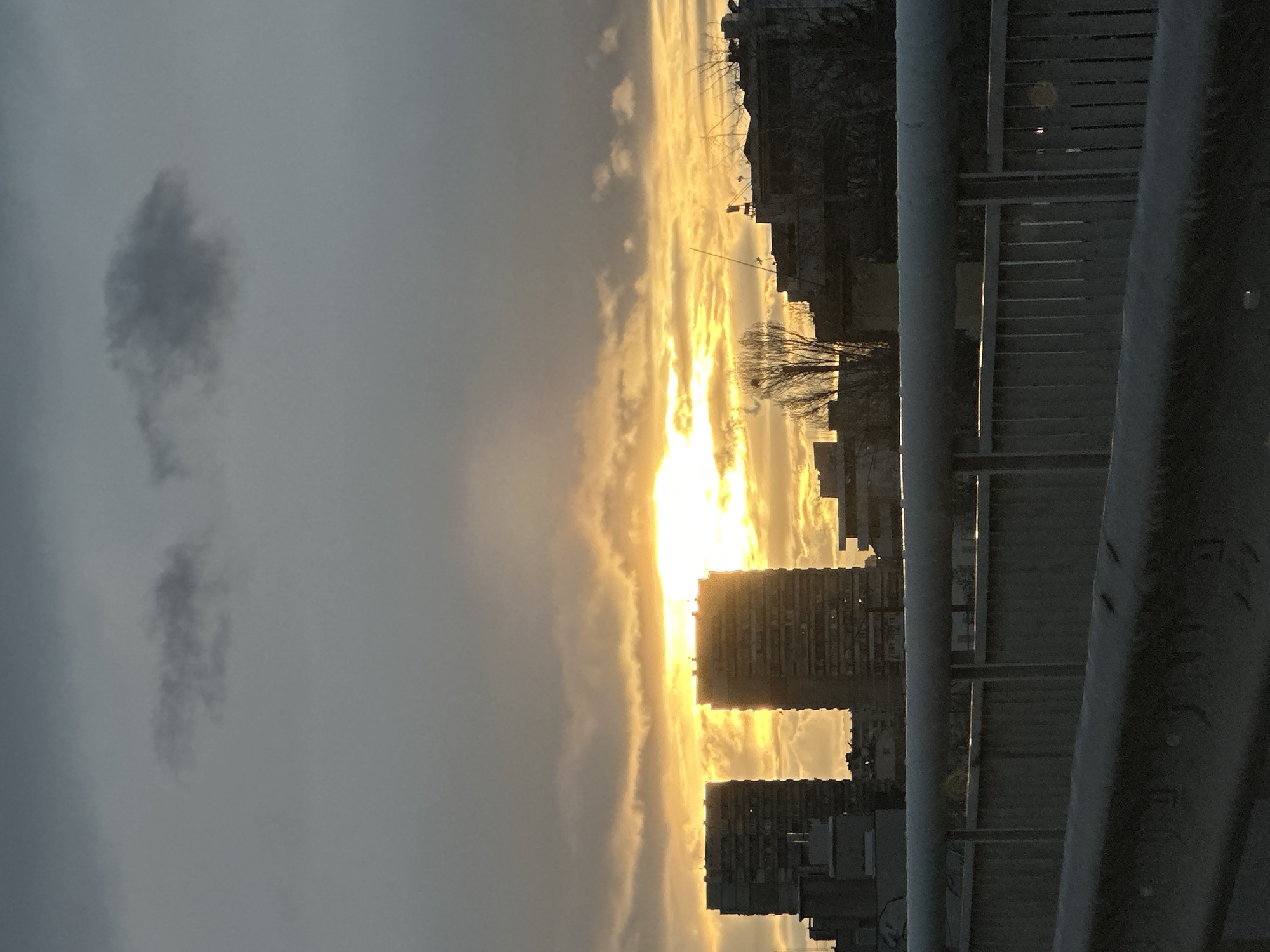 The image captures a view from an elevated position in Sofia, Bulgaria, possibly from a bridge or upper floor of a building. The scene features a vivid sunset with streaks of gold and light filtering through the clouds and between buildings. Silhouetted buildings stand against the backdrop of the setting sun. The photograph's right side features a metallic guardrail, adding a sense of depth and indicating the vantage point. A solitary cloud is visible in the sky. Overall, the picture showcases an urban landscape at dusk.