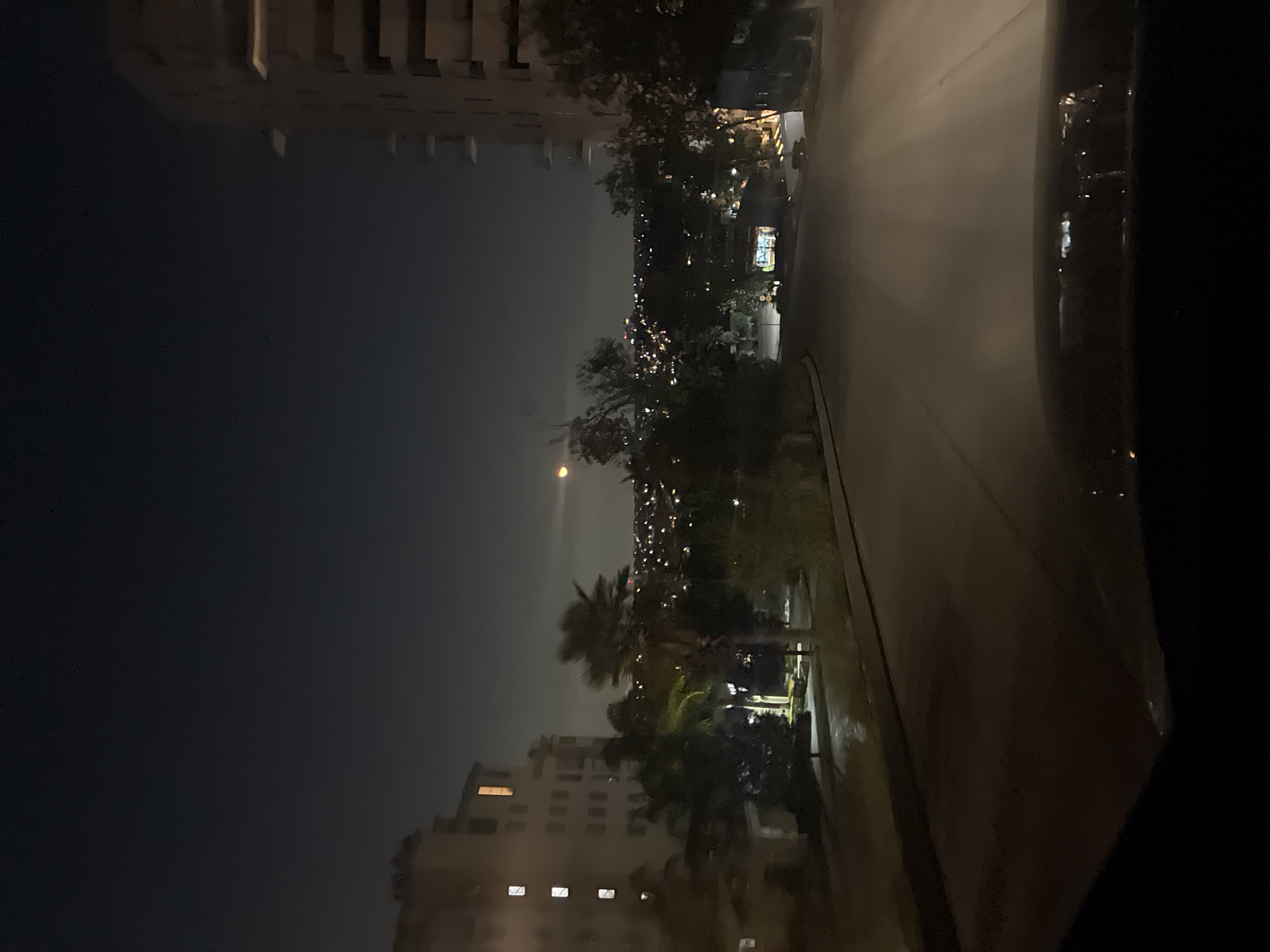 The image shows a nighttime urban scene in Huixquilucan de Degollado, Mexico, from the perspective of inside a vehicle. A road is visible, illuminated by streetlights, alongside a line of trees and vegetation. Buildings are present on both sides of the frame. The upper portion of the frame displays a cityscape with numerous lights indicating residential or commercial activity. The sky above is dark.