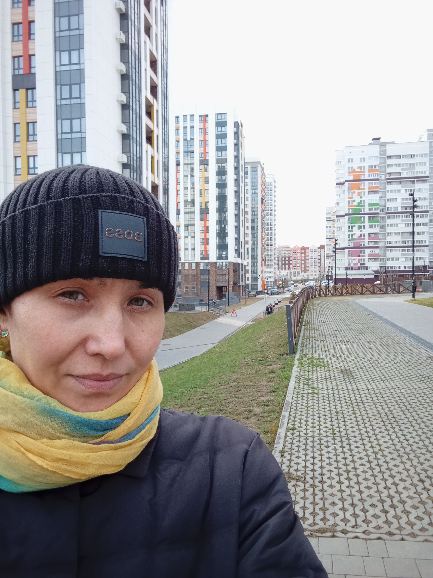 A person stands in front of buildings on a street in Izhevsk, Russia. The person wears a black beanie with a "BOSS" logo, a yellow and teal scarf, and a dark jacket. High-rise apartment buildings dominate the background, with a paved walkway to the right. A low wooden fence separates the walkway from a grassy area. Cars and other buildings can be seen further down the street. The sky is overcast.