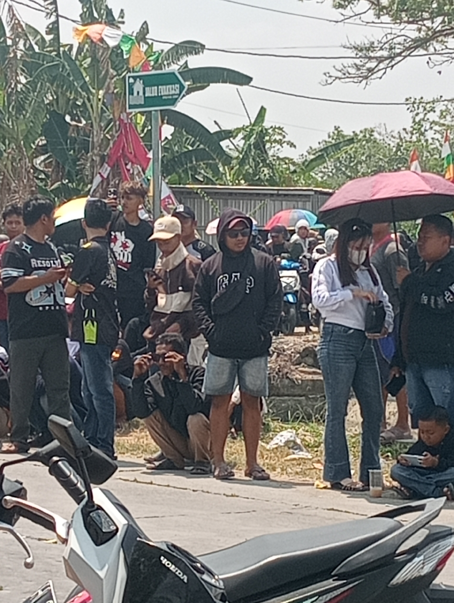 In the city of Doreng, Indonesia, a crowd gathers outdoors, possibly for an event or gathering. A green directional sign, with text in Indonesian and an icon of a building, points to the right, indicating an "evacuation route." The sign is adorned with colorful flags, and more flags are visible on nearby trees, suggesting a festive atmosphere.
Several individuals are present, with many looking at their phones or interacting with each other. Some are holding umbrellas for shade, including a prominent red umbrella in the foreground. A motorcycle is parked in the lower part of the frame. The ground appears to be a paved road or pathway, with some debris and foliage visible. The weather seems to be bright and sunny.