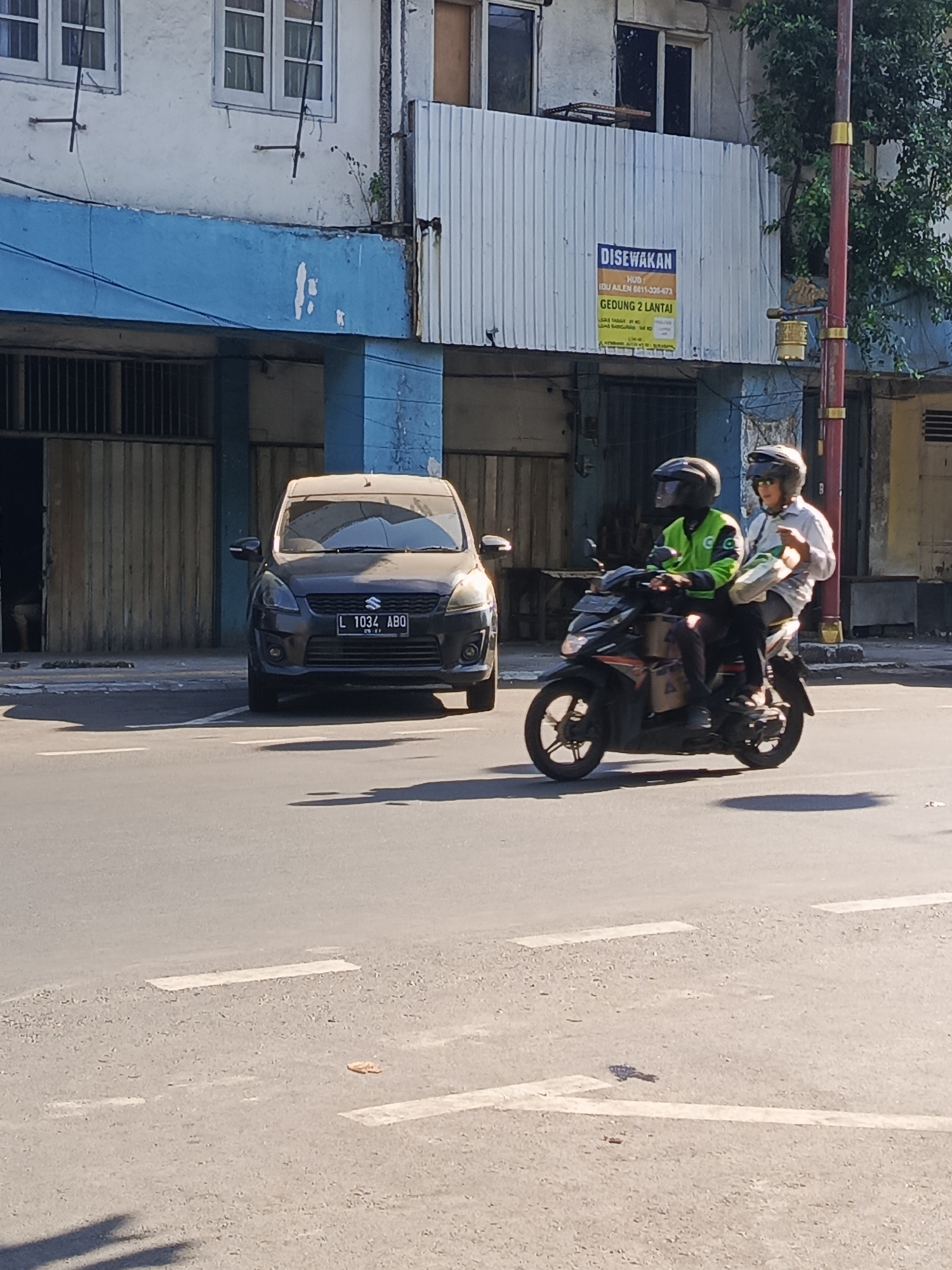 In the city of Surabaya, Indonesia, on a sunny day, a dark grey Suzuki Ertiga is parked on the street. The license plate reads "L 1034 AB0". To the right of the car, two individuals are riding a motorcycle. The driver wears a bright green jacket, and the passenger wears a white long-sleeved shirt. Both are wearing helmets. The motorcycle is black with orange accents and has delivery boxes attached to the sides. Behind the vehicles, a building with a blue facade and white corrugated metal siding is visible. A sign on the building reads "DISEWAKAN" which means "FOR RENT" in Indonesian, and details information about renting a two-story building. The scene takes place on a paved road with white lane markings.