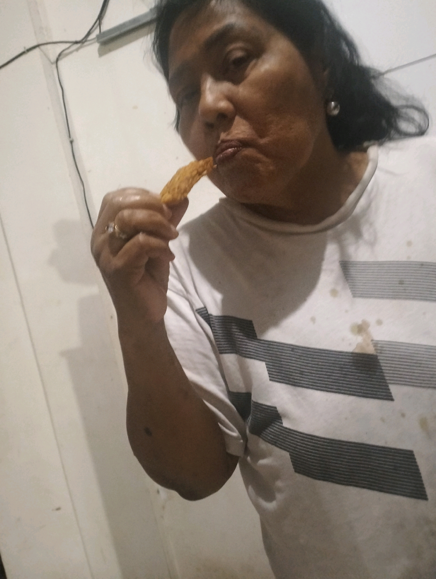 A middle-aged Indonesian woman in a white t-shirt with gray horizontal stripes eats a fried snack in Klecorejo, Indonesia. Her right hand, adorned with a ring, holds the snack to her mouth. The t-shirt has a few food stains. The background is a plain, light-colored wall with some wires and conduits visible.