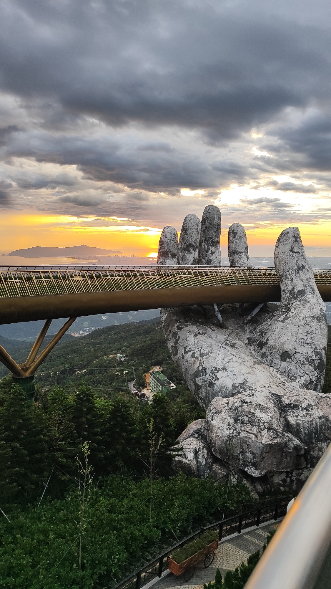 This image captures the Golden Bridge in Đà Nẵng, Vietnam, during a sunset. The iconic pedestrian bridge, supported by two massive stone hands, is seen extending across a mountainous landscape. The sun is setting over the horizon, casting a warm glow on the sky and the distant city skyline. Lush green trees and vegetation cover the mountainside, with cable cars visible in the background, suggesting a tourist attraction. The stone hands appear weathered and textured, adding to the dramatic visual of the bridge. The sky is filled with dramatic, cloudy formations, with patches of sunlight breaking through. In the foreground, a paved walkway with decorative stone patterns and a wooden cart filled with greenery lead the eye towards the main subject.