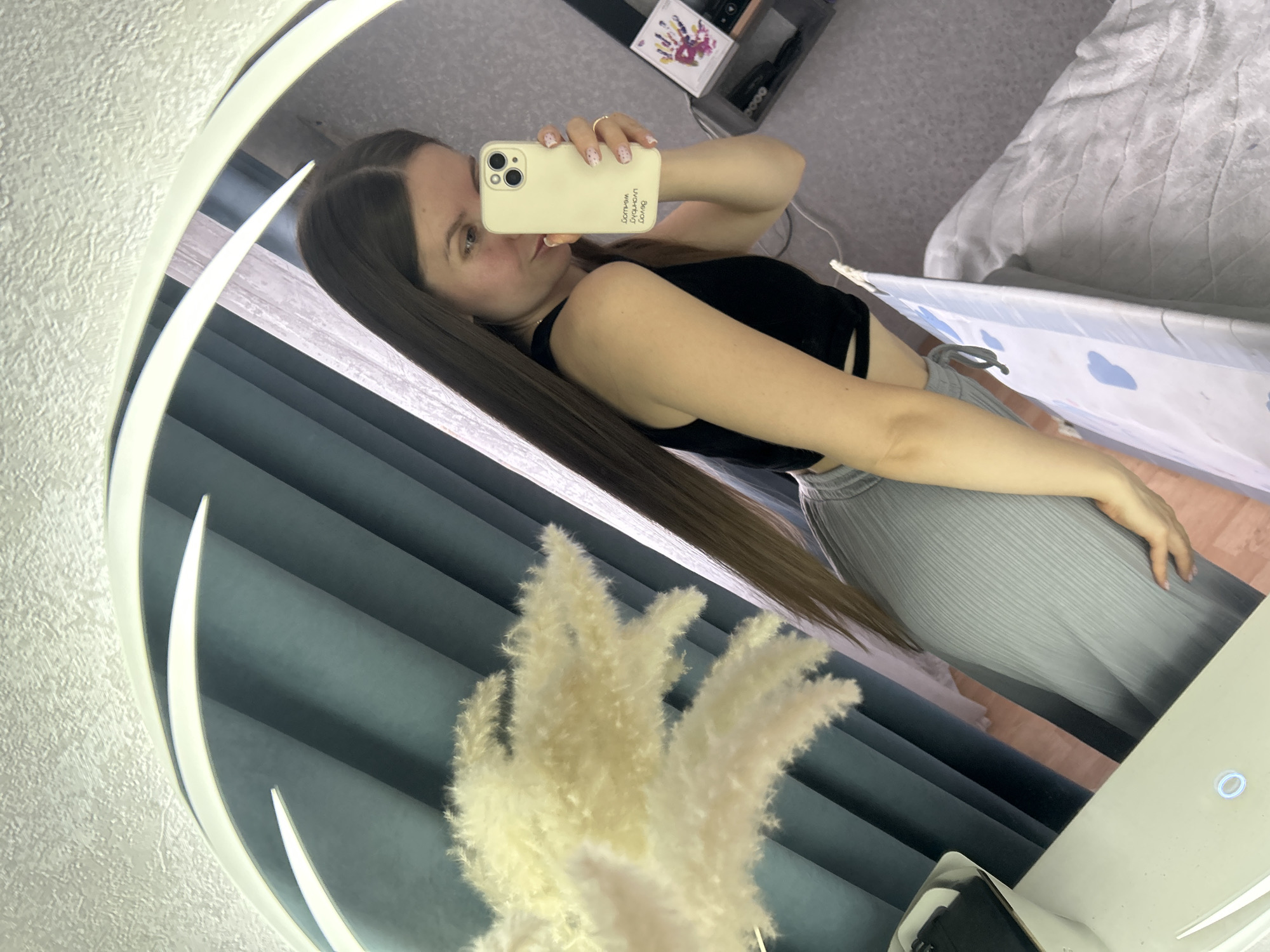 A young woman is captured in a mirror selfie taken indoors in what appears to be a residence in Klin, Russia. She has long, straight dark brown hair and is wearing a black crop top and light grey pleated pants. She holds a pale yellow smartphone with an iPhone-like camera setup, her hand adorned with a ring on her finger, as she poses for the photo. Her fingernails have a light-colored manicure with small dark dots.
The mirror itself is large and round, featuring bright, illuminated accents around its edge. In the reflection, the background shows a textured light grey wall and dark grey or blue blinds or curtains. A fluffy, light-colored pampas grass decoration is visible in the lower-left foreground, reflected in the mirror. To the right, a bed covered with a light grey blanket and a white and blue baby playpen or cot are visible. A shelf with various items, including a card with colorful handprints, can also be seen in the reflection.
On the back of the woman's phone case, visible text in Russian is reversed due to the mirror image. It appears to read "Осчастливь меня" (Make me happy) and "люблю" (love). The setting suggests a casual, indoor environment, possibly a bedroom or living area.