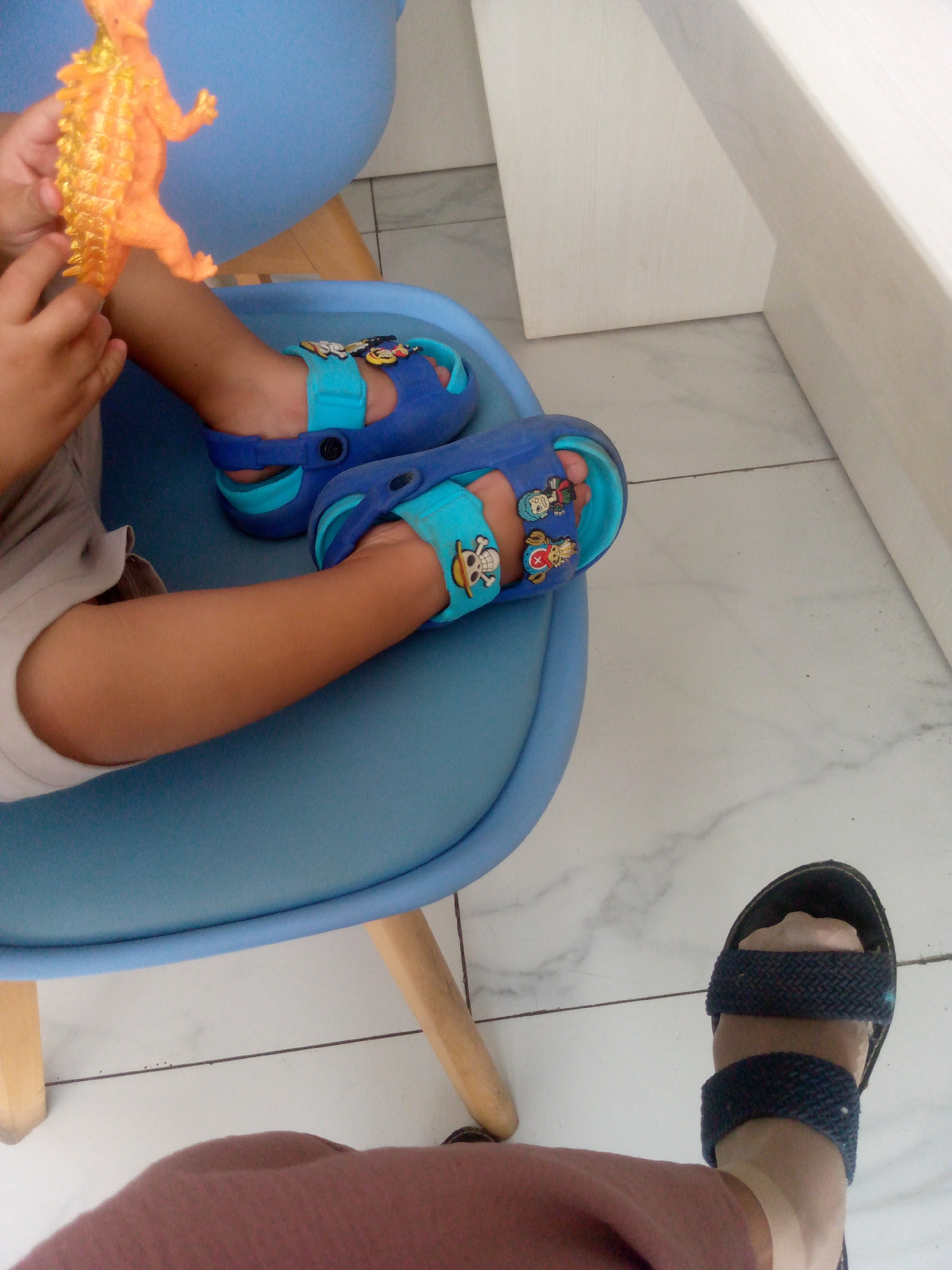 A child's bare legs and feet are visible, clad in blue clogs with cartoon character embellishments. The child is holding an orange dinosaur toy. The child is seated on a blue chair, which has wooden legs. The flooring is tiled and appears to be white with grey veining. In the foreground, an adult's legs and feet are visible, wearing dark sandals and brown pants. The scene appears to be indoors.