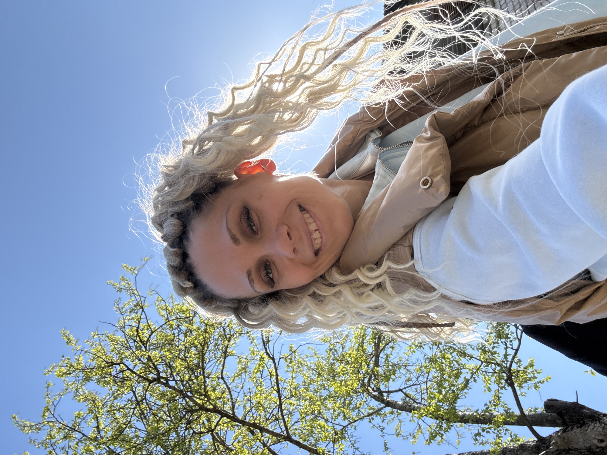 A low-angle selfie captures a woman with blonde, braided hair and a wide smile. She is wearing a light-colored sweatshirt and a brown jacket. The background consists of a bright blue sky and tree branches with budding green leaves. It cannot be confirmed that this location is in the city of Rusanovo, Russia.