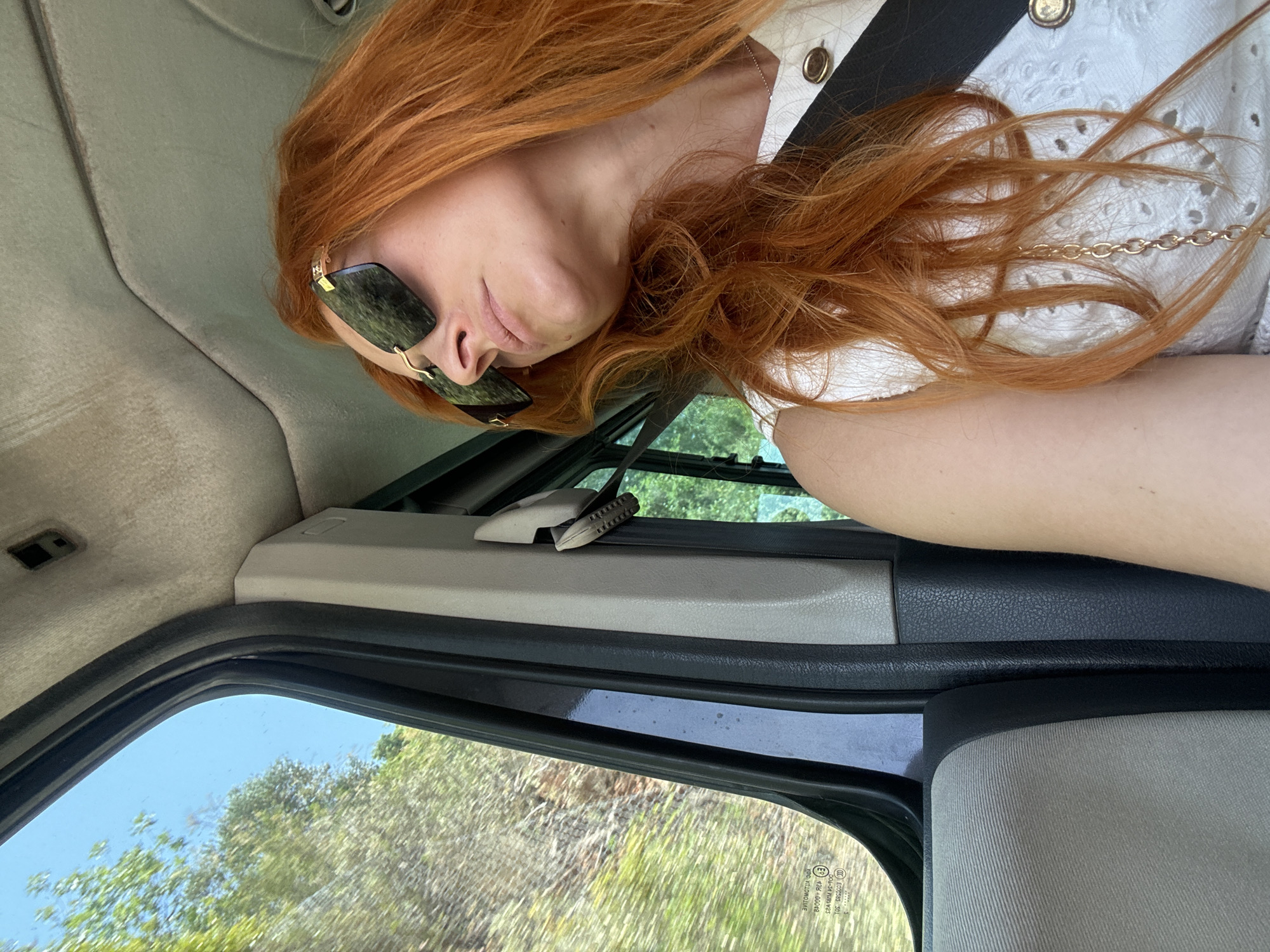 A young woman with long, wavy, ginger hair is resting her head on her arm inside a vehicle. She is wearing large, square, dark-tinted sunglasses with thin gold frames. Her left hand is resting on her cheek, and her right arm is visible in the lower right portion of the frame. She is wearing a white top with eyelet detailing and gold buttons. The interior of the vehicle appears to be grey and black. Through the open window, lush green trees and vegetation are visible against a bright blue sky, suggesting it is daytime and the weather is clear. The scene suggests a leisurely drive through a natural landscape. The image is rotated 90 degrees counterclockwise. No text is clearly visible.