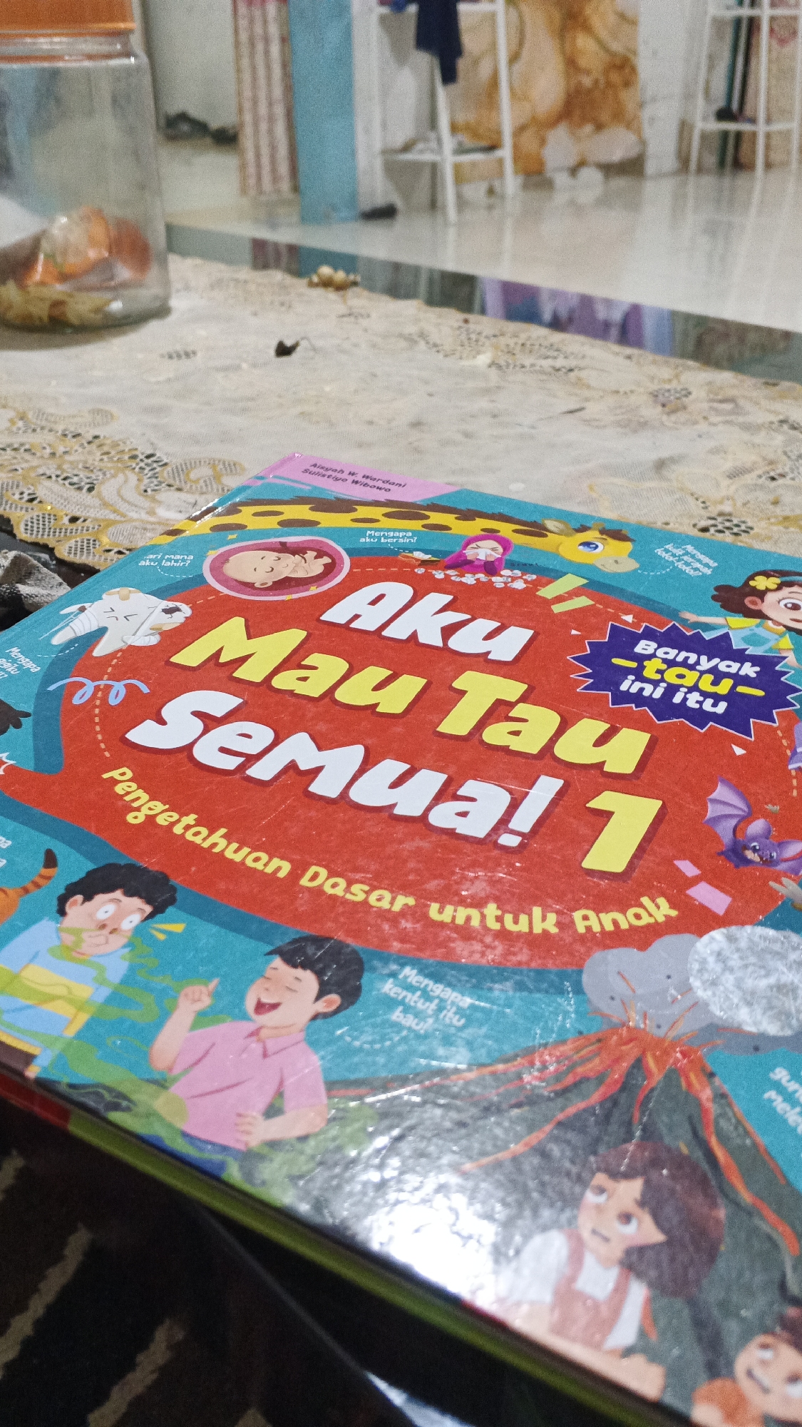 The image shows a close-up of a children's book titled "Aku Mau Tau Semua! 1" with the subtitle "Pengetahuan Dasar untuk Anak". The book cover is red and features cartoon illustrations of children, a baby, a giraffe, a bat, and a volcano. Some text in Indonesian is visible on the cover, including "Banyak -tau- ini itu". The book is lying on a patterned fabric surface. In the background, out of focus, is a clear glass jar with orange contents and a blurry background suggesting an indoor setting with furniture. The scene is not in the city of Kampung Rempak, Indonesia; it is an indoor scene focused on a book.