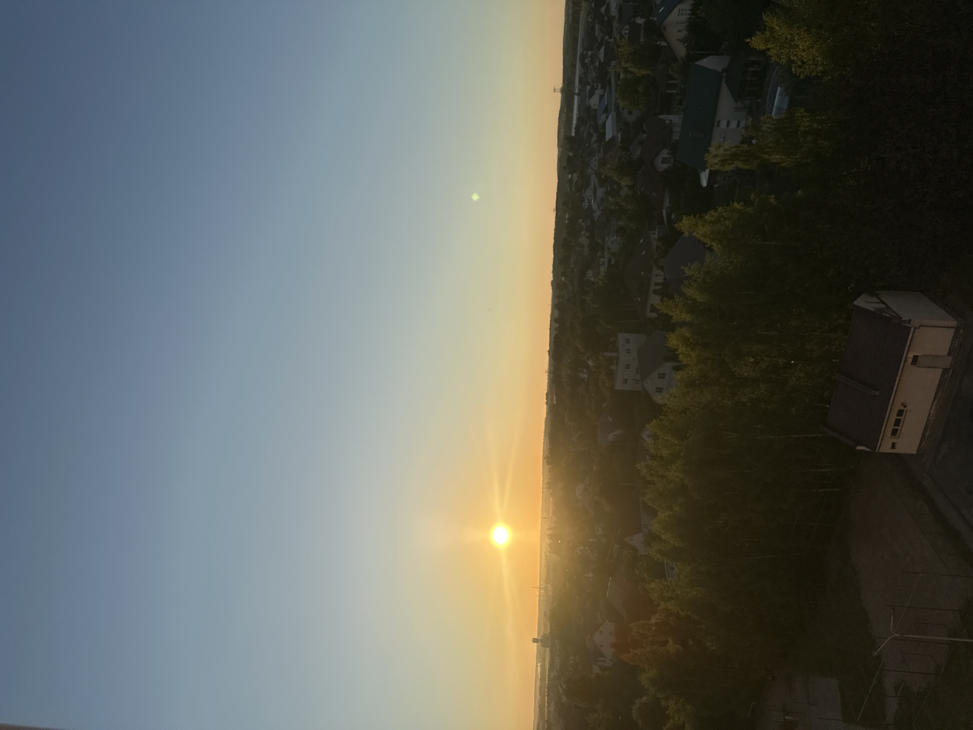 The image depicts a panoramic view of a suburban area during sunset, likely in the vicinity of Шчомысліцкі сельскі Савет, Belarus. The sky is a gradient of pale blue at the top, transitioning to a warm orange and yellow near the horizon where the sun is setting. The sun itself is a bright, distinct orb, casting a radiant glow across the scene.
Below the sky, a dense expanse of trees, predominantly leafy and green, forms a natural barrier separating the foreground from the residential area. This foliage is partially illuminated by the setting sun, creating areas of both light and shadow.
Scattered throughout the landscape are numerous residential buildings, characterized by their varied roof colors, including dark grey, green, and white. These houses are nestled amongst the trees and on gently rolling terrain, suggesting a well-established residential community. In the distance, faint outlines of industrial structures or water towers can be seen, hinting at the proximity of more developed areas or infrastructure.
In the immediate foreground, a darker, more enclosed area is visible, featuring a sports court with visible lines and fencing. Adjacent to this court is a small, rectangular building with a dark roof and light-colored walls, exhibiting a simple, utilitarian design with a few small windows. Another similar structure is partially visible to the right, indicating the presence of additional facilities or outbuildings. The overall impression is one of a tranquil, residential setting bathed in the warm, atmospheric light of a sunset.