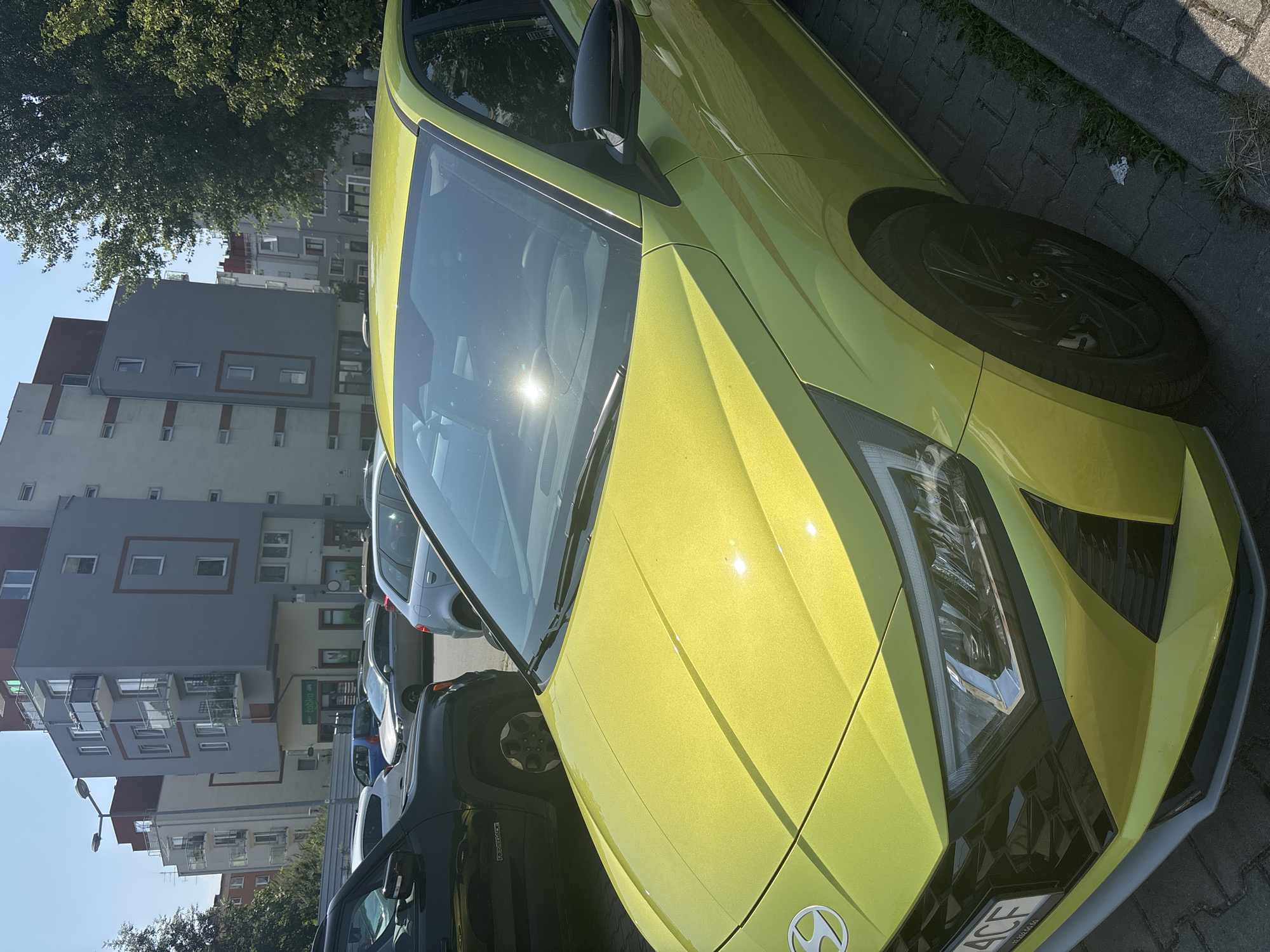 In Krakow, Poland, on a sunny day, a bright yellow Hyundai i20 is parked on a cobblestone street in front of apartment buildings. The car is facing right, with its front visible. The foreground is dominated by the car's hood and front bumper. In the background, several multi-story apartment buildings with balconies and windows are visible. Trees line the street, and other vehicles are parked nearby, including a black Jeep. The overall scene suggests a typical urban residential area.