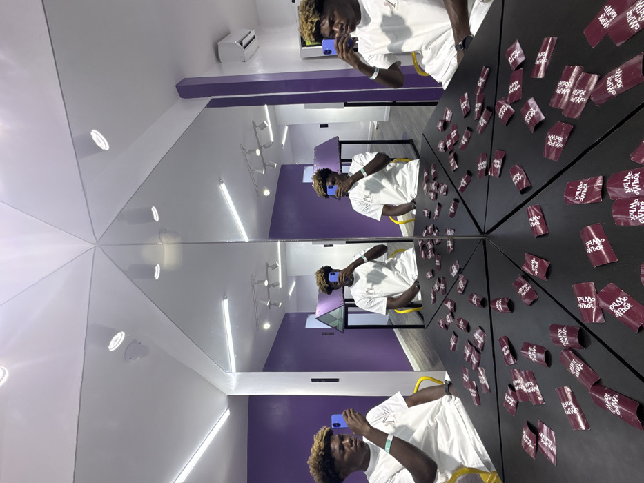 The image depicts a young male positioned within a highly reflective, mirrored room located in Jakande, Nigeria. The central subject, a male with dark skin and short, coily blonde hair, is wearing a white t-shirt with a subtle logo, a light blue wristband on his left wrist, and a dark watch on his right. He is seated on a yellow chair, actively holding a blue-cased smartphone in his left hand, pointed towards himself, indicating he is taking a selfie or recording.
The room's design incorporates extensive mirrors, creating multiple reflections of the individual, appearing as numerous identical figures seated around a central structure. This central structure is a black, reflective, multi-faceted table or platform, which is densely covered with numerous small, curled, burgundy-colored paper strips. These strips display white text, repeatedly showing "Wha? What?".
The environment features vibrant purple walls, a white ceiling fitted with multiple recessed spotlights and linear fluorescent light fixtures, and a light-colored floor. An air conditioning unit is visible on the upper right wall. In the mirrored reflections, further sections of the purple-walled room and a triangular geometric frame are discernible in the background.