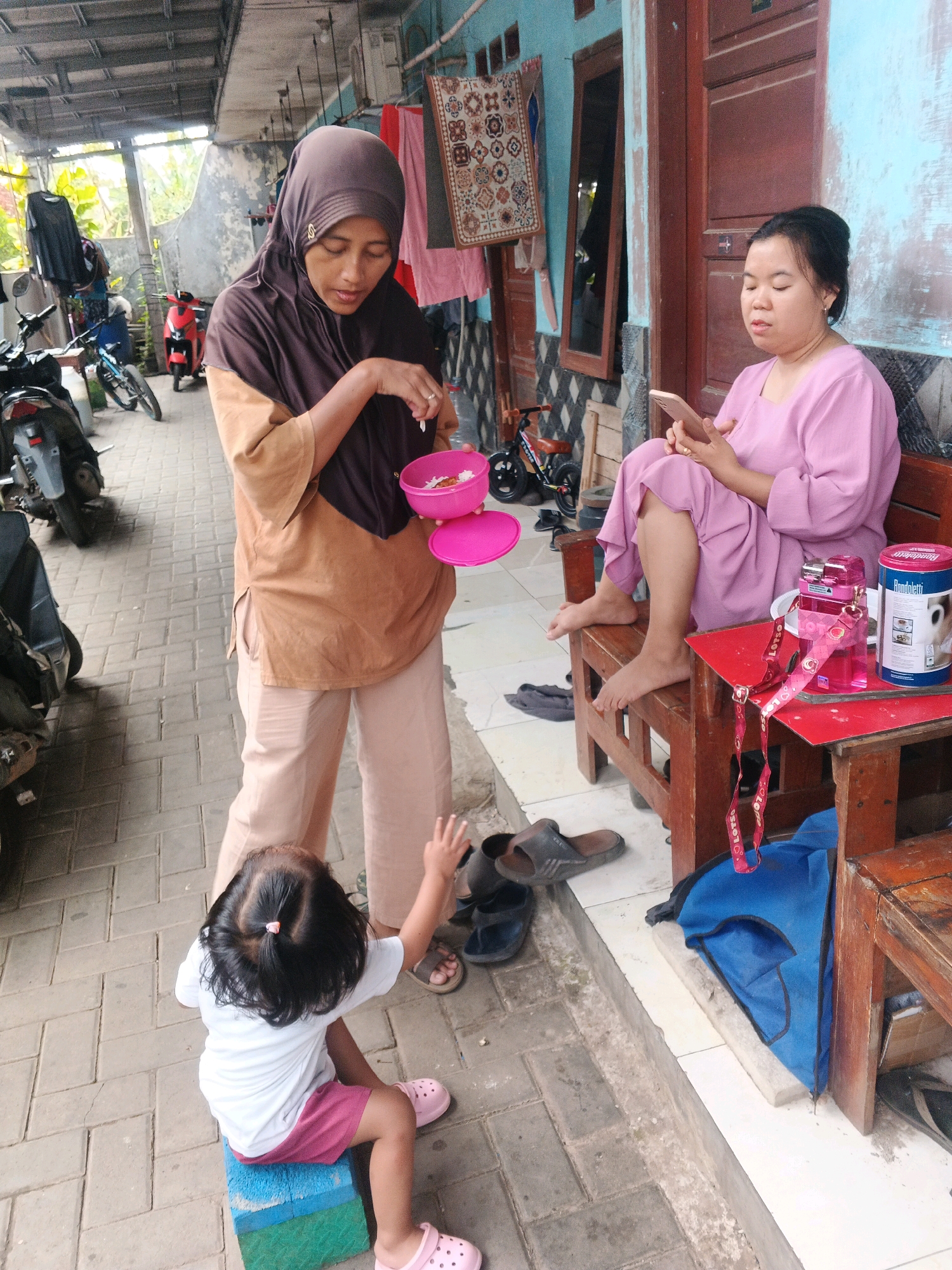 In Kadu, Indonesia, a street scene unfolds under a covered outdoor area. A woman wearing a brown hijab and a tan tunic, holding a pink bowl of rice, stands over a young girl. The girl, wearing a white t-shirt and pink shorts, sits on a colorful block, her back to the camera, looking up and reaching towards the woman. Beside them, another woman in a pink outfit is seated on a wooden bench, engrossed in her phone. A red table next to the seated woman holds a pink water bottle and a cylindrical container. The environment shows a tiled walkway, motorcycles parked nearby, and a wooden door with blue walls in the background.