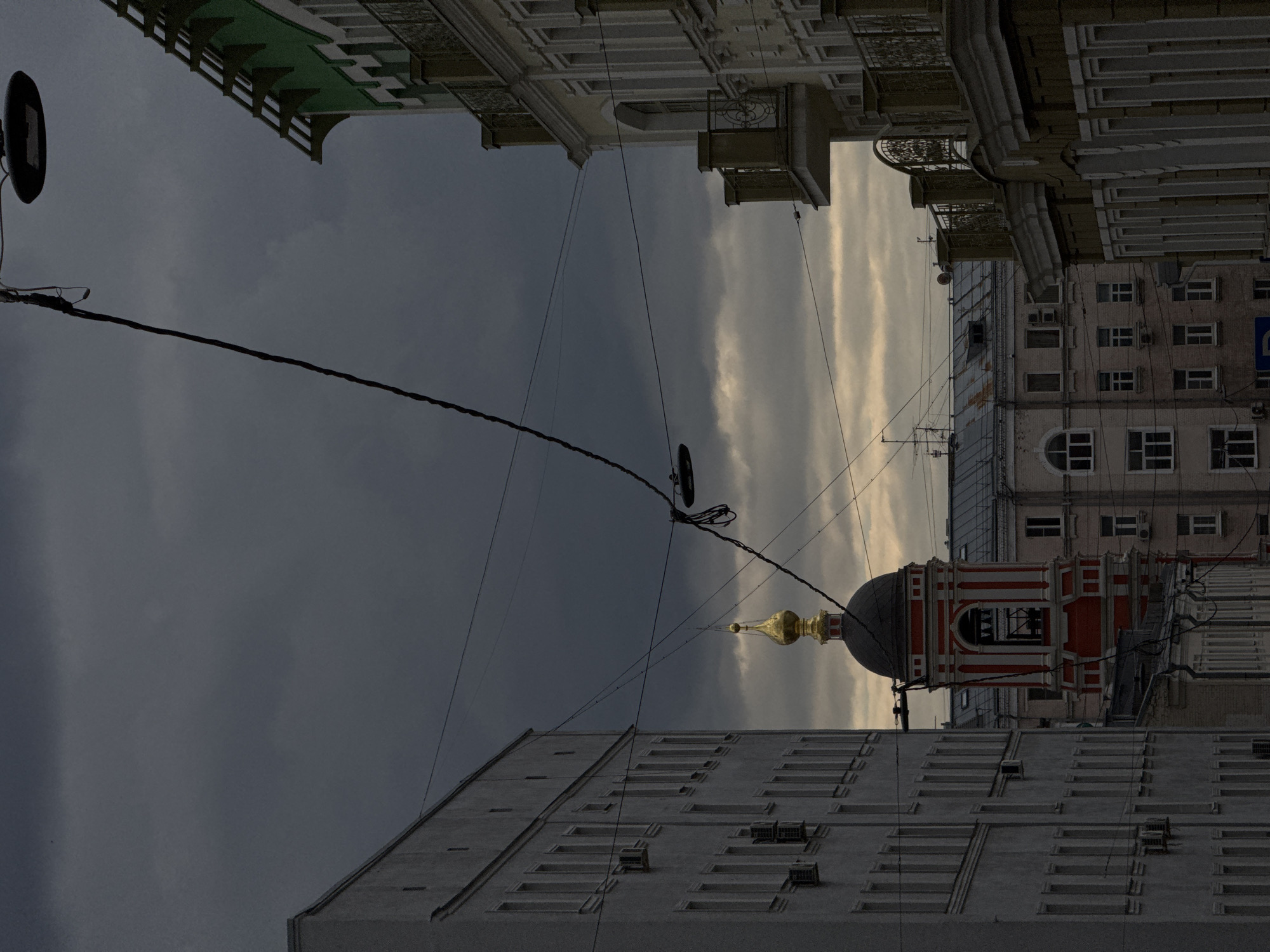 The image displays an urban landscape in Moscow, Russia, under a dramatic, overcast sky, likely during late afternoon or early evening. The scene is dominated by several buildings and a crisscross of overhead utility wires.
In the foreground, on the left, stands a tall, grey multi-story building with numerous regularly spaced rectangular windows. To its right, a distinctive red and white church-like structure is visible, featuring a dark dome topped with a bright golden cupola and spire, characteristic of Russian Orthodox architecture.
Above and to the right of the church, an ornate, classical-style building with intricate facades, decorative balconies, and cornices adds to the architectural variety. Further in the background, other residential or commercial buildings can be seen, some equipped with air conditioning units and exhibiting various window designs.
The sky is heavily clouded, with dark, brooding grey clouds occupying the left and central parts of the frame. Towards the right, the clouds appear lighter and more stratified, showing hints of pale yellow or orange, suggesting either a break in the cloud cover or the light of a setting sun. A dark streetlamp is visible on the far left, connected to some of the numerous overhead wires that traverse the scene. No people are visible. A small blue sign is faintly discernible on a distant building, but any text on it is unreadable. The overall mood conveyed by the heavy sky and varied architecture is one of quiet urban contemplation.