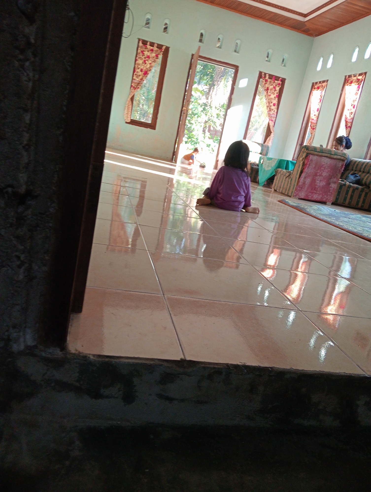 A young child in a purple shirt and pants sits on a tiled floor in a brightly lit room in Campago Barat, Indonesia. The child is facing away from the camera, looking towards an open doorway that leads outside to a lush, green area. Sunlight streams into the room through the open door and several tall, narrow windows, creating reflections on the shiny floor. The room appears to be a living area with furniture, including sofas and what looks like a table covered with a blue cloth. The walls are painted a light blue, and the ceiling is made of wood. In the distance, another person can be seen near a sofa. The overall impression is a peaceful domestic scene bathed in natural light.