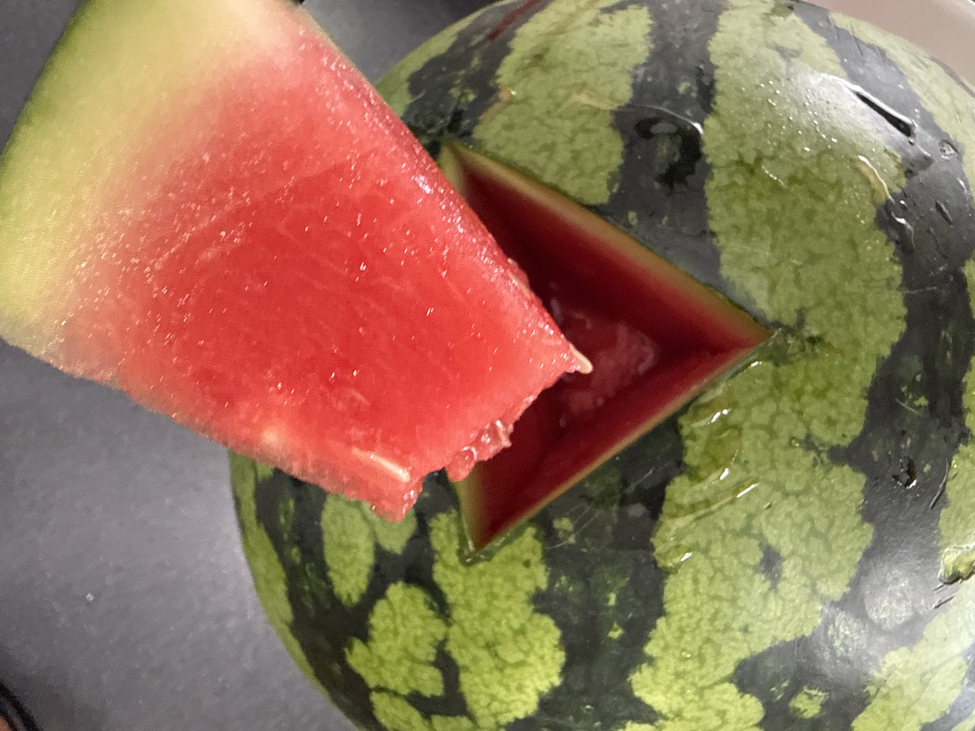 A close-up, overhead shot shows a triangular wedge of watermelon being extracted from a larger, whole watermelon. The extracted wedge is bright red with a light green rind, showing a gradient from deep red to yellow-green. The main body of the watermelon is visible, with its characteristic dark green and light green striped rind. A triangular opening has been cut into the top of the watermelon, revealing the deep red flesh within. The scene is shot from above, with a dark, slightly reflective surface beneath the watermelon, possibly a countertop. The lighting is bright, highlighting the textures and colors of the fruit. No people are visible in the frame. The location is the city of Marum, Netherlands.