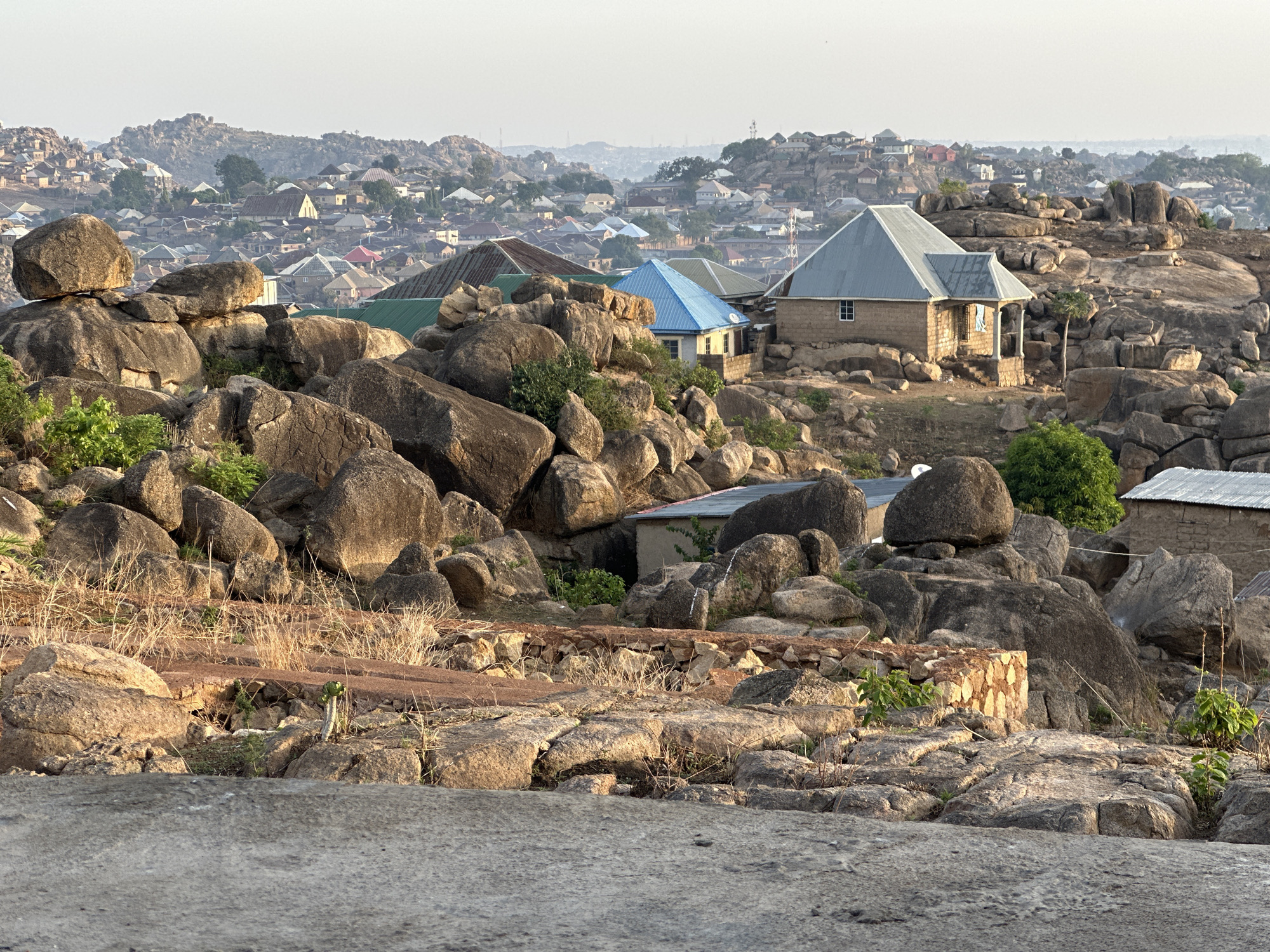 The image depicts a landscape in Jos, Nigeria, characterized by scattered houses and large boulders. The foreground features a rocky terrain with dry grass and small plants. A stone structure, possibly a wall, is visible amidst the rocks. Several houses with corrugated metal roofs, some painted blue or green, are nestled among the boulders. In the background, a dense settlement of houses extends across the hilly landscape, fading into a hazy horizon. The sky is pale, indicating either early morning or late afternoon.