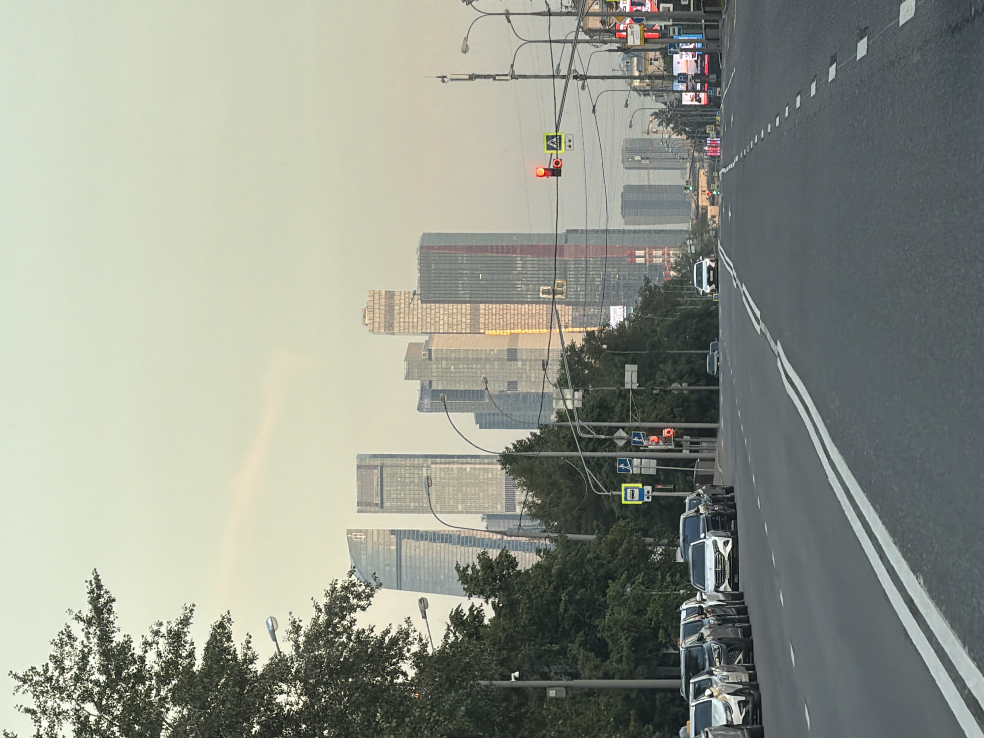 The image depicts a wide, multi-lane asphalt road extending into the distance under a pale, hazy sky, characteristic of Moscow Oblast, specifically referencing Khimki, Russia. In the far background, a cluster of modern skyscrapers, consistent with the Moscow International Business Center (Moscow City), rises against the skyline. Two prominent skyscrapers on the left feature glass facades, one with a curved profile and a red emblem near its peak, the other rectangular. Further right, other skyscrapers display varying architectural elements, including one with a striking red vertical stripe.
In the mid-ground, the road is divided by solid and dashed white lines. On the left side of the road, a line of parked or stopped vehicles, including several dark SUVs/sedans and a white Mercedes-Benz sedan (license plate "X321XH 750", indicating Moscow Oblast), is positioned next to a dense row of green trees forming a median or park area. A white taxi with a yellow roof light is at the front of this line of vehicles. On the right side, the main traffic lanes show a single white vehicle (possibly an SUV) in the distance, approaching a green traffic signal.
Numerous grey lampposts with streetlights line both sides of the road. On the left, above the parked cars and trees, a blue bus stop sign and a red pedestrian traffic light are visible. On the right side, a red vehicle traffic light hangs over the road, accompanied by a yellow pedestrian crossing sign. Utility poles with multiple wires and cellular antennae are also present on the right. Further down the road on the right, large digital billboards display illuminated content. No individuals are discernible in the scene. The overall lighting suggests either early morning or late afternoon with diffuse sunlight.
