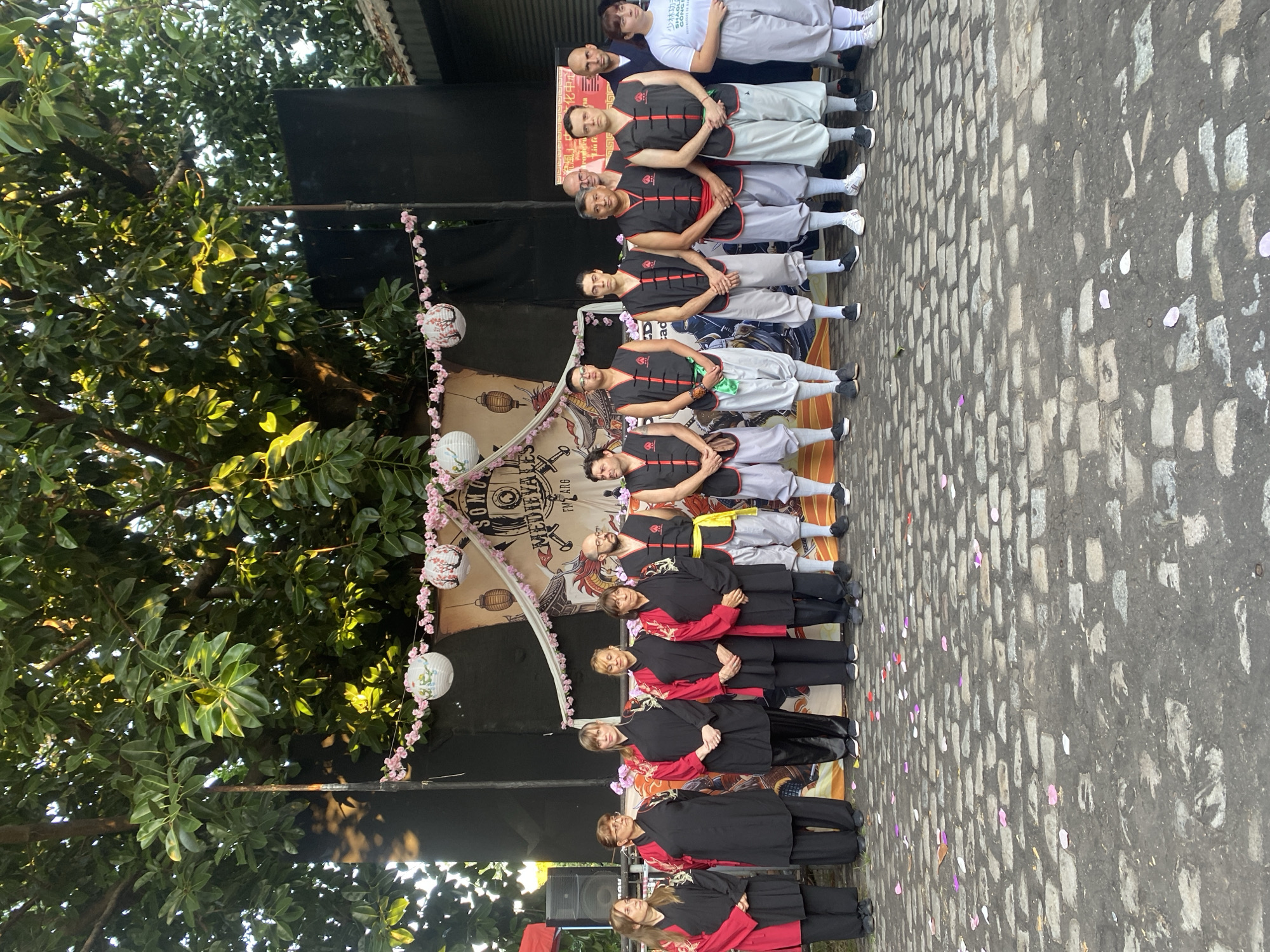 The image shows a group of people lined up on a cobblestone street in what appears to be Buenos Aires, Argentina. The group is arranged on a stage or platform, dressed in various martial arts attire. Some wear black and red uniforms, while others wear black vests with red stripes over light grey pants. There are trees on the left side of the frame, and decorative lanterns and streamers are visible around the stage area. The background includes a banner with writing in what appears to be medieval text. Some of the individuals are smiling, and the overall impression is that they are participating in a performance or public event.
