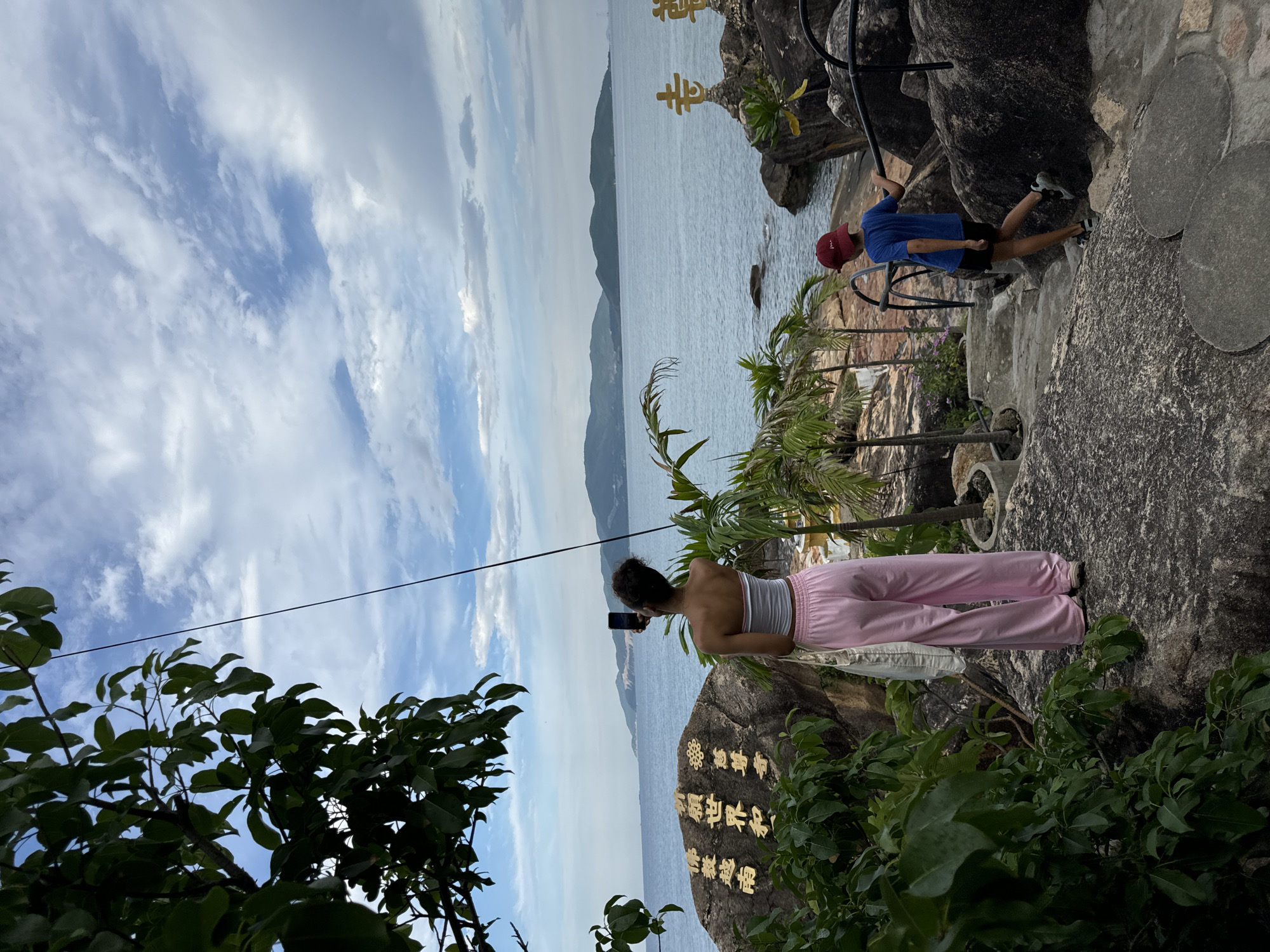In Nha Trang, Vietnam, two people are on a rocky coast. A young woman, wearing a white tube top and pink wide-leg pants, stands on a large rock, holding a smartphone to take a picture of the ocean. Beside her on the rock are large, gold inscriptions. To her right, a young boy in a blue t-shirt and red baseball cap walks down a stone path, holding onto a black metal railing. The ocean stretches out to the horizon, with distant islands visible under a cloudy sky. Lush green foliage frames the scene, with palm trees and large rocks dotting the landscape.