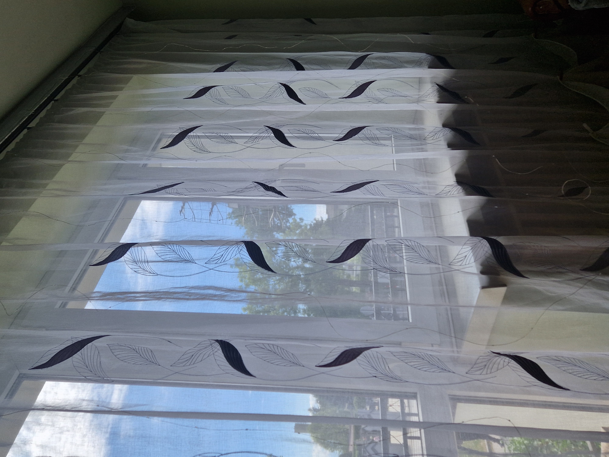 A static, eye-level indoor photograph depicts a translucent white sheer curtain set covering a multi-pane window during daytime. The curtains feature a repeating vertical pattern consisting of dark, solid, elongated leaf shapes interspersed with outlined leaf designs showing internal venation, rendered in a thin, dark line. The fabric is gathered, forming vertical folds, and is suspended from a dark curtain rod visible along the top edge. Through the curtains and window, a bright blue sky with scattered white clouds is visible in the upper left quadrant. A dense landscape of green trees occupies the middle ground and background. In the distant left, a slender, dark, cylindrical tower-like structure is discernible. Below the treeline, vague architectural elements of distant buildings and a horizontal railing are subtly visible. A light-colored wall (possibly pale green or beige) frames the right side of the window opening. A flat, light-colored surface, consistent with a window sill or a piece of furniture, is present below the window. The overall illumination is bright, characteristic of clear daytime conditions.