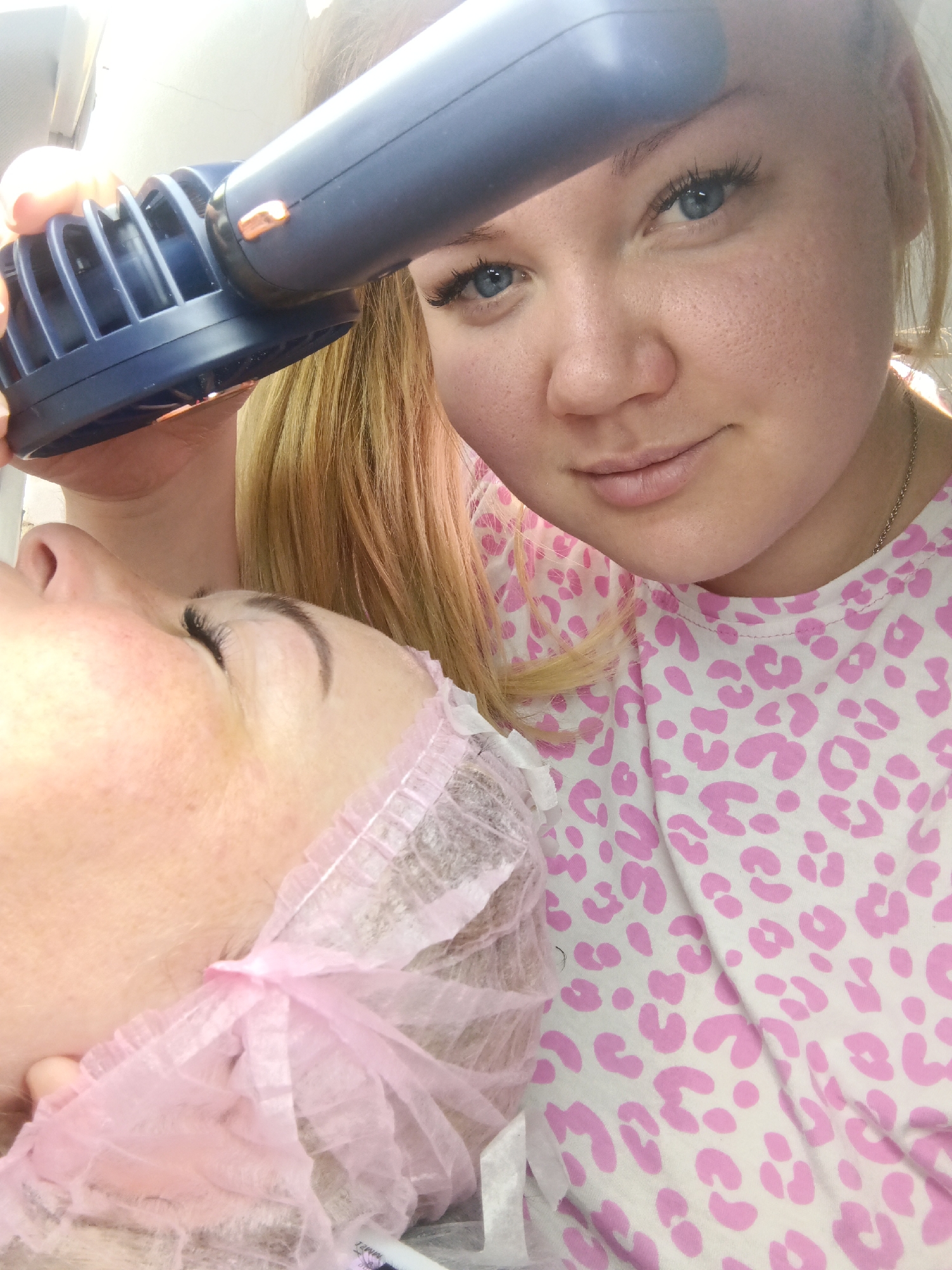 A medium shot depicts a woman applying beauty treatment to another woman who is lying down. The woman applying the treatment is holding a dark blue device with an attachment on the side, positioned over the face of the client. The client has her eyes closed and is wearing a pink hair net. The woman applying the treatment is looking at the camera and wearing a white shirt with a pink leopard print. The setting appears to be a beauty salon or a similar professional setting. The location given is Фоки, Russia.