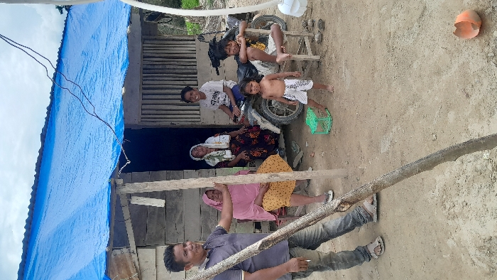 The image captures a group of people, likely a family, gathered outdoors in front of a simple dwelling. The scene is set on uneven, light brown dirt ground.
In the foreground, slightly to the left, a man wearing a dark grey/blue collared shirt and blue jeans stands, looking towards the camera with a faint smile. He is holding a long, slender wooden pole that extends diagonally upwards and to the right across the frame. He is wearing sandals.
Behind the man, and under a rudimentary shelter, two women are seated. One woman, positioned closer to the center, wears a patterned dress and a white head covering. She is looking towards the camera. Next to her, to the right, another woman is seated, wearing a pink garment and possibly a yellow patterned skirt, also smiling towards the camera.
To the right of the seated women, a dark-colored motorcycle is parked on the dirt. A young person, possibly a teenager, in a white t-shirt and shorts, is seated on the motorcycle, smiling. Standing beside the motorcycle, another child, shirtless and wearing patterned shorts, also looks towards the camera. Behind the motorcycle, a smaller child in a white top and dark shorts is sitting on a low wooden stool, smiling.
The dwelling behind the group appears to be constructed from rough wooden planks. A large blue tarp serves as a roof or covering for part of the structure, visible on the left side of the image. Some exposed electrical wires are seen hanging from the upper left portion of the structure.
Other objects visible in the scene include a green plastic basket or crate near the motorcycle and an orange, broken helmet or bowl lying on the dirt ground in the upper right area of the image (when correctly oriented). The sky in the upper left appears bright, indicating it is daytime, possibly with overcast conditions or bright sunlight. The overall mood conveyed by the smiling faces is one of casual gathering and warmth. No discernible text is visible in the image.