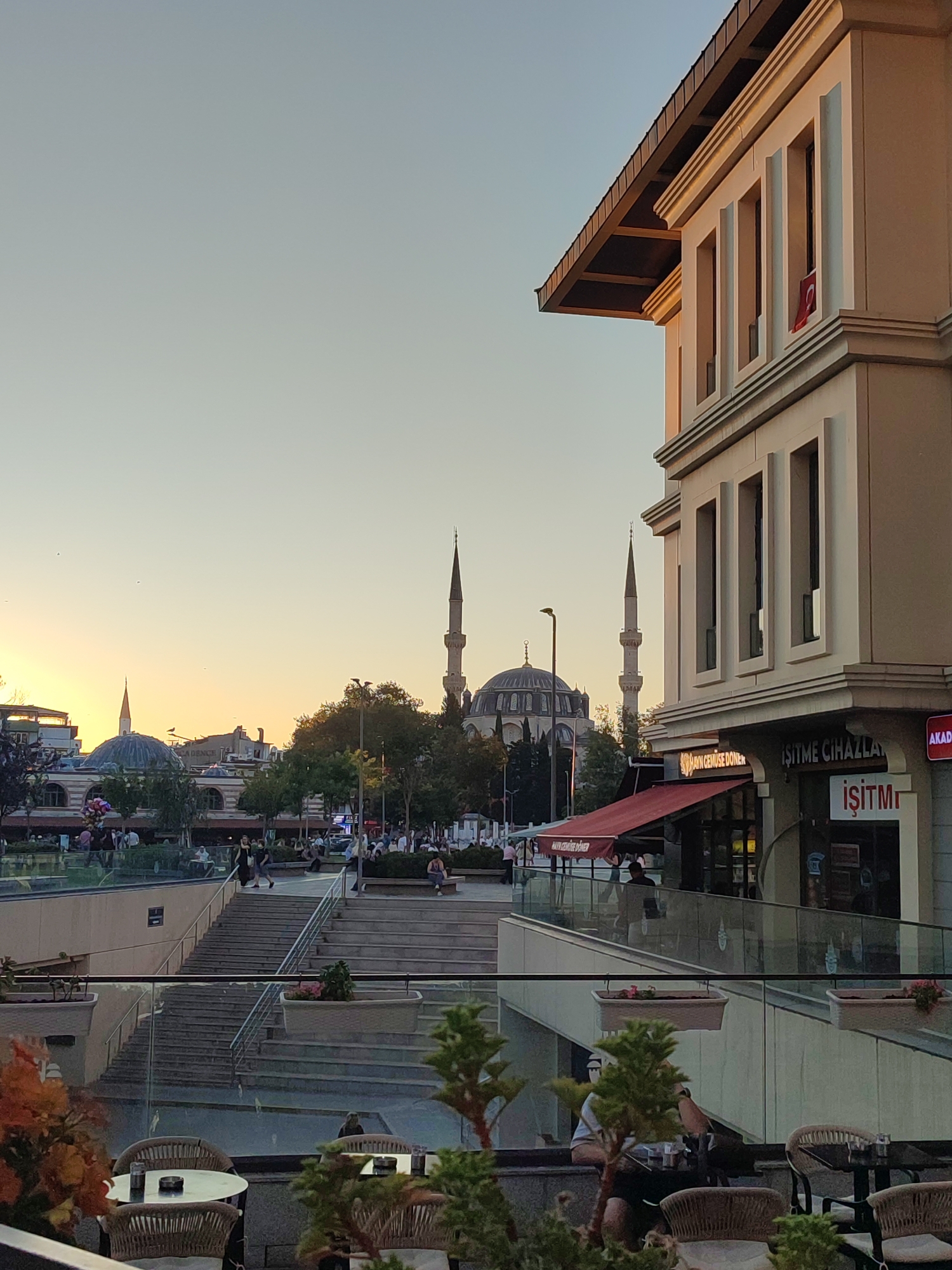 The image captures a street scene in Üsküdar, Turkey, likely during the golden hour due to the warm lighting. In the foreground, a modern building with a sloped roof and multiple windows is visible on the right. A Turkish flag hangs from one of the windows. Below the building, there is a commercial area with awnings and storefronts, including signs that appear to be in Turkish. A glass railing separates this area from a lower level where people are seated at tables with chairs, some with ashtrays and glasses on them, suggesting an outdoor seating area of a cafe or restaurant.
Beyond the modern building, the iconic silhouette of the Blue Mosque (Sultan Ahmed Mosque) and its minarets are prominent in the background, partially obscured by trees and other buildings. The sky above is a clear gradient of pale blue and yellow, typical of sunset or sunrise.
In the mid-ground, a large set of outdoor stairs leads down to a lower level. Several people are visible on and around these stairs, some walking, others sitting. Further in the distance, more people are gathered in an open area, with balloons visible adding a touch of color. The overall scene depicts a bustling urban environment with historical landmarks integrated into a modern cityscape.