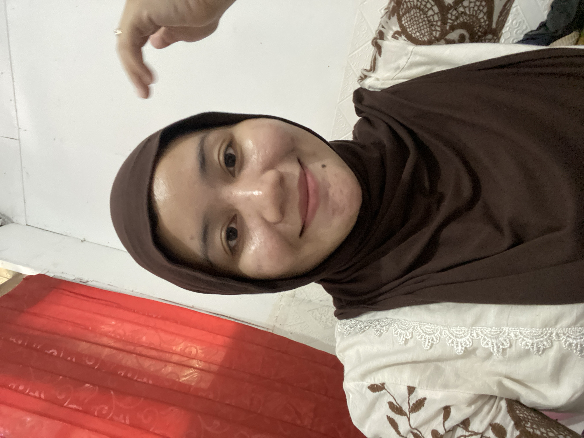 A young Indonesian woman with fair skin and dark hair is captured in a close-up, waist-up selfie. She is wearing a dark brown hijab and a white shirt adorned with brown floral embroidery and white lace trim. Her skin appears slightly oily, and she has a subtle smile. Her right hand is raised, and a ring is visible on her index finger. In the background, a red curtain with a patterned texture hangs on the left, and a white, textured wall is on the right. The lighting suggests an indoor setting, possibly a home in Banjarbaru, Indonesia.