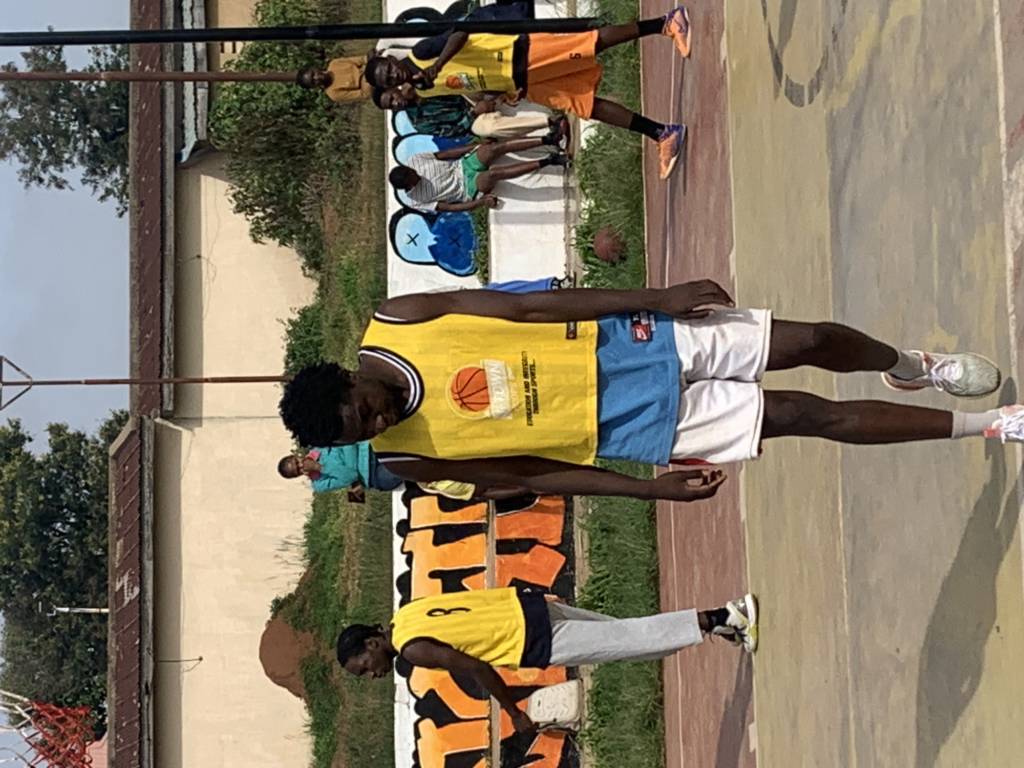 In Gura Top, Nigeria, a basketball game is underway on an outdoor court. The court surface is a mix of red and yellow asphalt, marked with white lines. A group of young men, dressed in basketball attire, are engaged in the game. One player in the foreground, wearing a yellow and blue jersey and white shorts, walks with his head down. To his left, another player in a yellow jersey and grey pants is seen from the side. In the background, several other individuals are observing the game, some seated on a low wall adorned with colorful graffiti. The court is enclosed by a chain-link fence, with a basketball hoop visible in the upper left corner. The environment appears to be in a semi-urban area, with buildings and vegetation in the background.