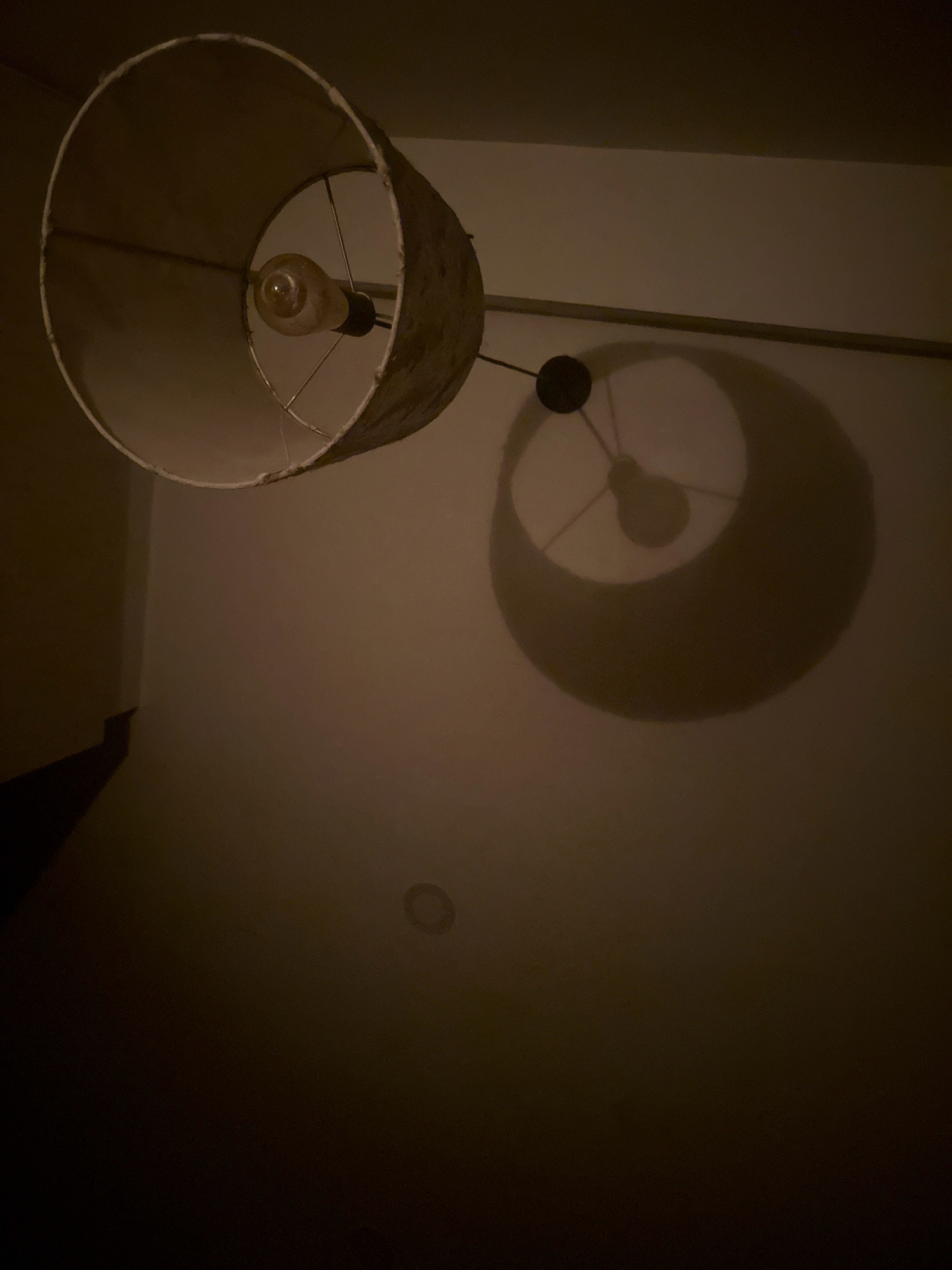 This media file shows a close-up, low-angle shot of a lampshade and its shadow cast on a wall. The lampshade is cylindrical with a textured, somewhat worn exterior and a lighter interior. A single light bulb is visible within the shade, emitting a warm, yellow glow. The shadow of the lampshade, with the filament of the bulb clearly defined, is projected onto the wall behind it, creating a circular shape with internal radial lines. A dark circular fixture, likely part of the lighting mechanism, is also visible. The background consists of a plain wall with a subtle gradient, suggesting ambient lighting. The overall mood is dark and somewhat moody, with the warm light of the bulb providing the main illumination. The scene appears to be indoors, given the lighting and the presence of a wall. No people, text, specific weather, or time of day cues are evident. The location context of Lalitpur, Nepal, provides a geographical reference but is not visually discernible from the image itself.
