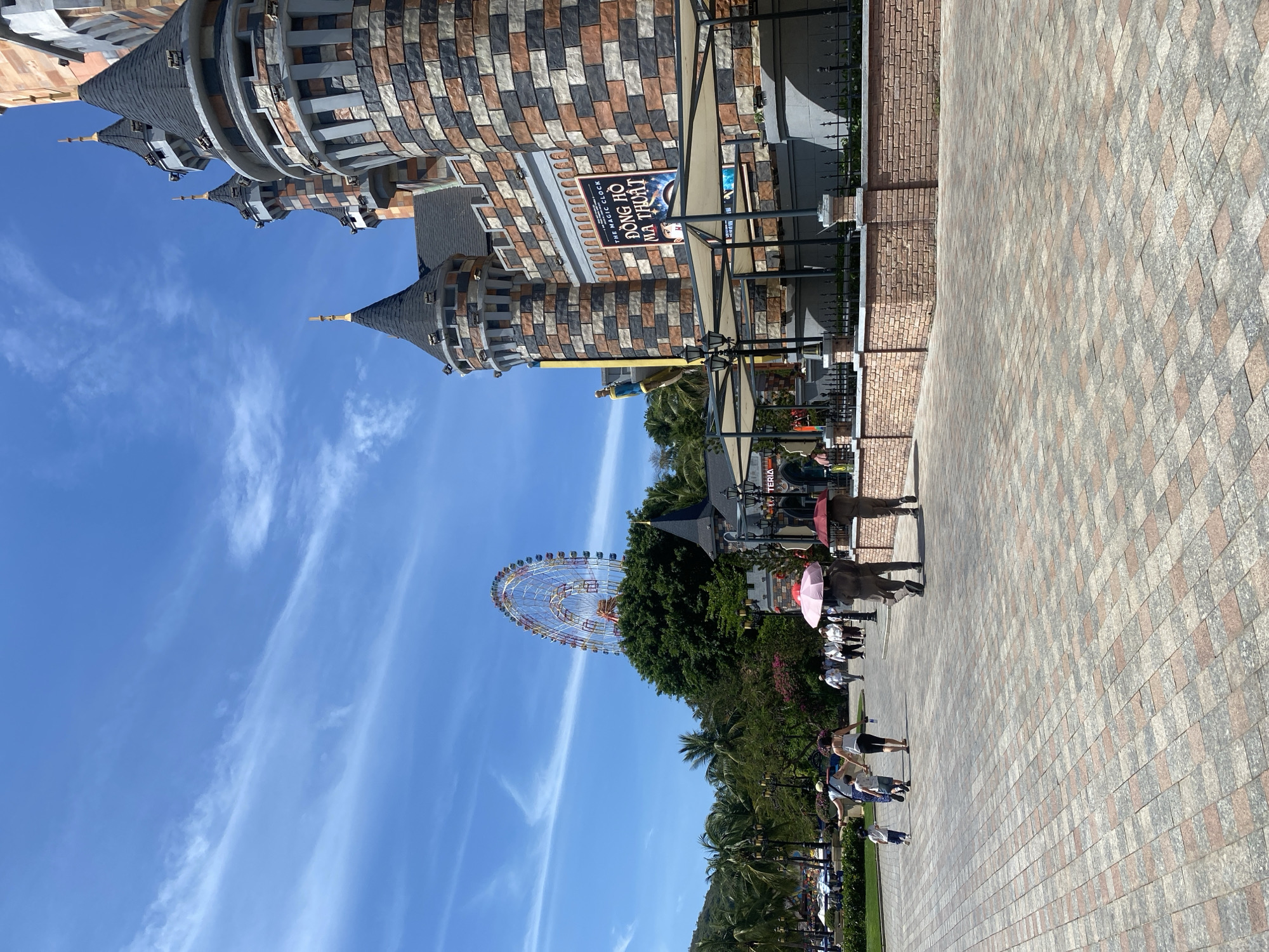 The image shows a street scene in an amusement park. On the left side of the image is a castle-like structure with several turrets and towers. A Ferris wheel is visible in the background. People are walking along the brick-paved path. Palm trees and other vegetation are on the left side of the path. A sign that says "Đồng Hồ Ma Thuật" can be seen. The sky is blue with some clouds.