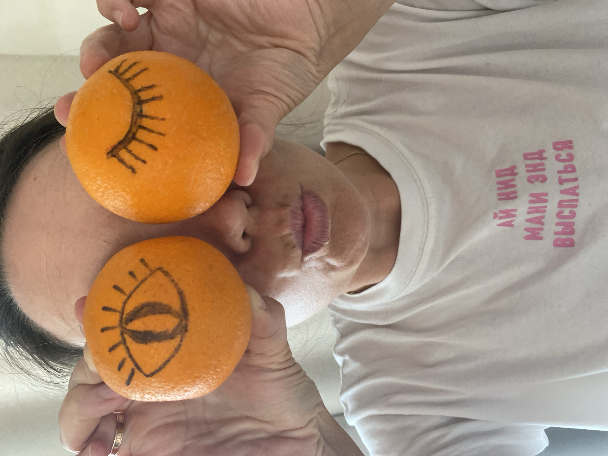 A person is holding two oranges, each with an eye drawn on it, in front of their face. The top orange has a closed eye drawn on it, while the bottom orange has an open eye drawn on it. The person is wearing a white t-shirt with pink text. The text on the shirt translates to "I need money and sleep". The image was taken indoors in Chaikovsky, Russia.