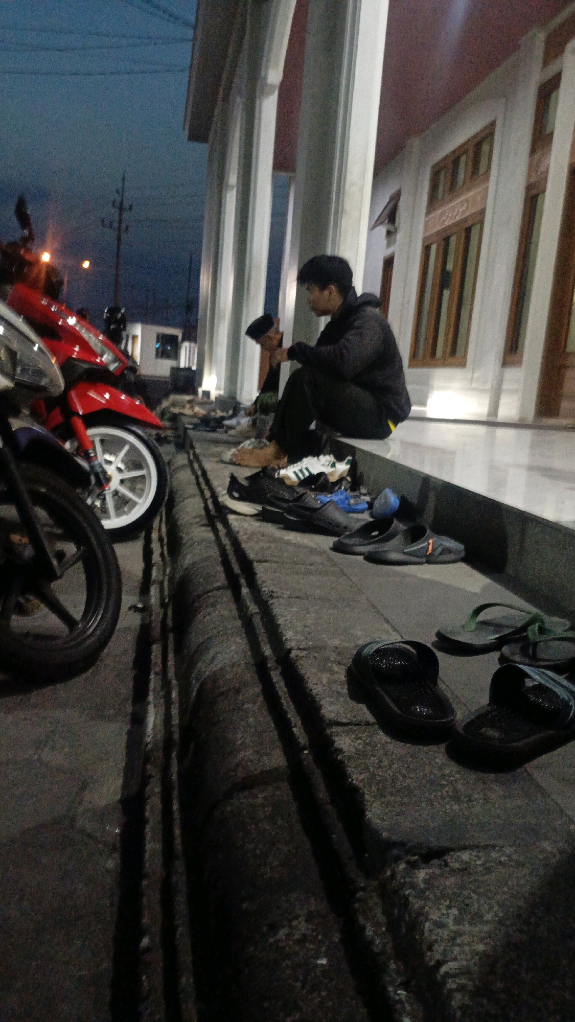 The image depicts a scene at dusk outside a building, likely a mosque given the architectural style with columns and windows, possibly in Ngaru Aru, Indonesia. Two young men are seated on a raised platform to the right, facing each other and appearing to be in conversation. One man, wearing a dark hooded jacket and dark pants, is looking down. The other, wearing a black cap and a light-colored garment, is looking towards the first man. In the foreground, several pairs of footwear are casually placed on the ground and the edge of the platform, including sandals and sneakers. Motorcycles are parked to the left, one predominantly red and white, and another partially visible in the foreground. The ground appears to be paved, with a noticeable drainage channel running along the edge of the platform. The lighting suggests it is either early morning or late evening, with ambient light from the sky and potentially artificial lights within or near the building. The overall atmosphere is relaxed and casual.