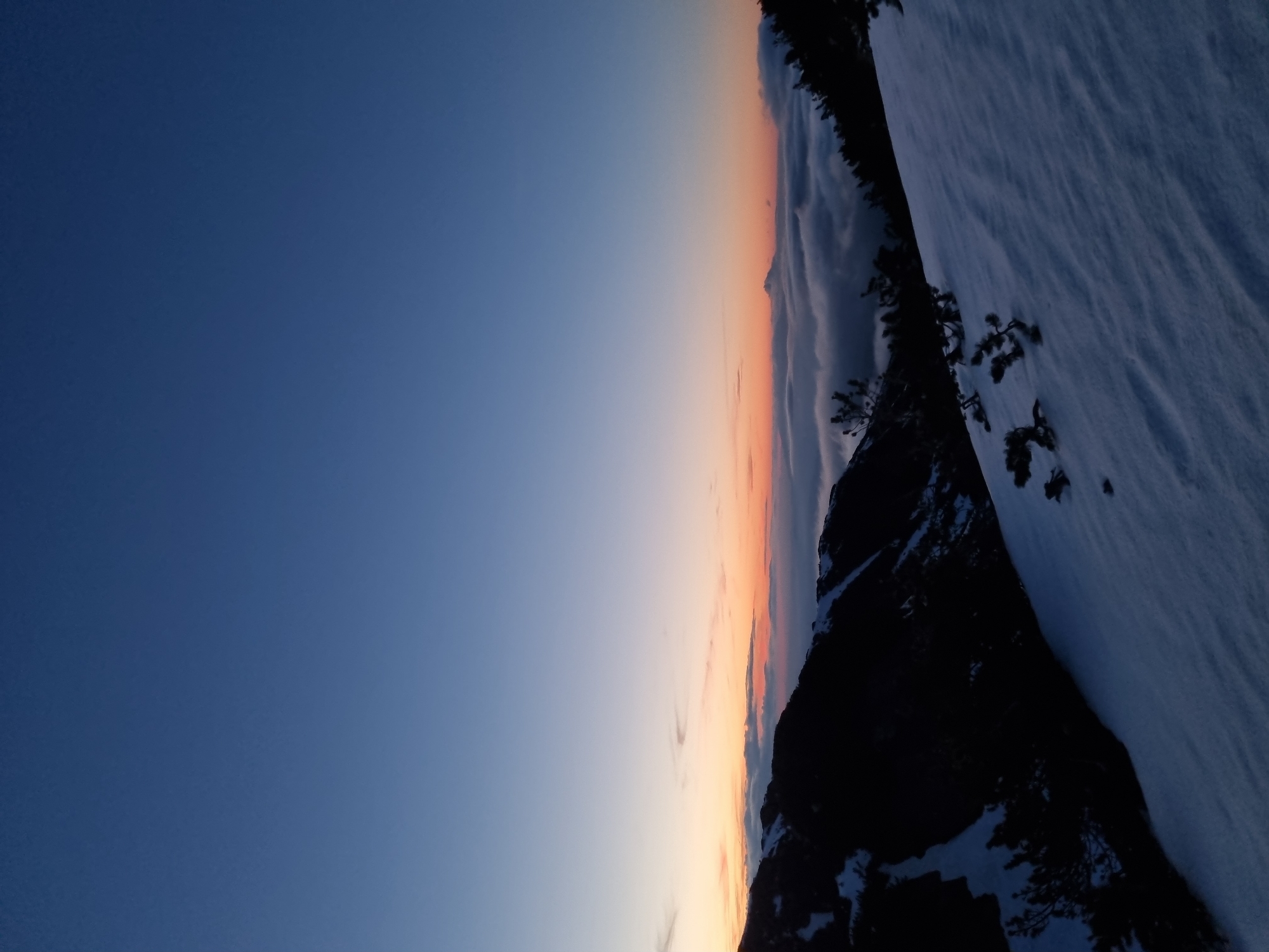 The image depicts a landscape with a dark blue sky transitioning into a gradient of orange and pink hues at the horizon. This horizon line is interrupted by a dark, silhouetted mountain range covered in snow. There are also some trees. The location is possibly near Govedartsi, Bulgaria, given the landscape, mountain, and sky features visible.