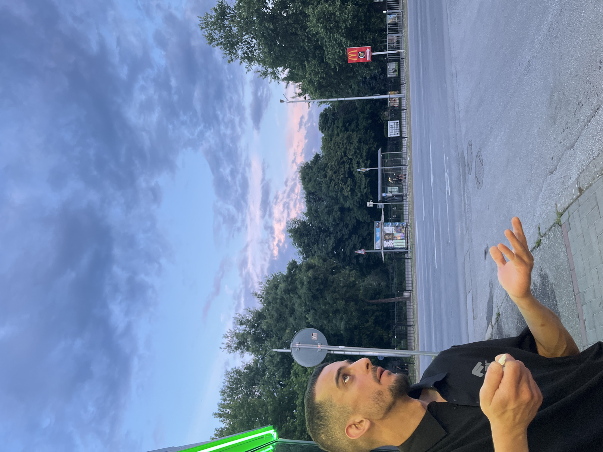 The image depicts a man in a black polo shirt standing on a sidewalk at dusk. He is looking upwards and to his left, with his right hand holding a small object and his left hand open, palm up. Behind him, a street is visible with traffic lanes and a bus stop shelter. Trees line the street, and in the distance, a McDonald's sign is illuminated. The sky is cloudy with hues of pink and blue, indicating either sunrise or sunset. The man's location is on a street corner in Plovdiv, Bulgaria, likely in an urban area with public transportation access.