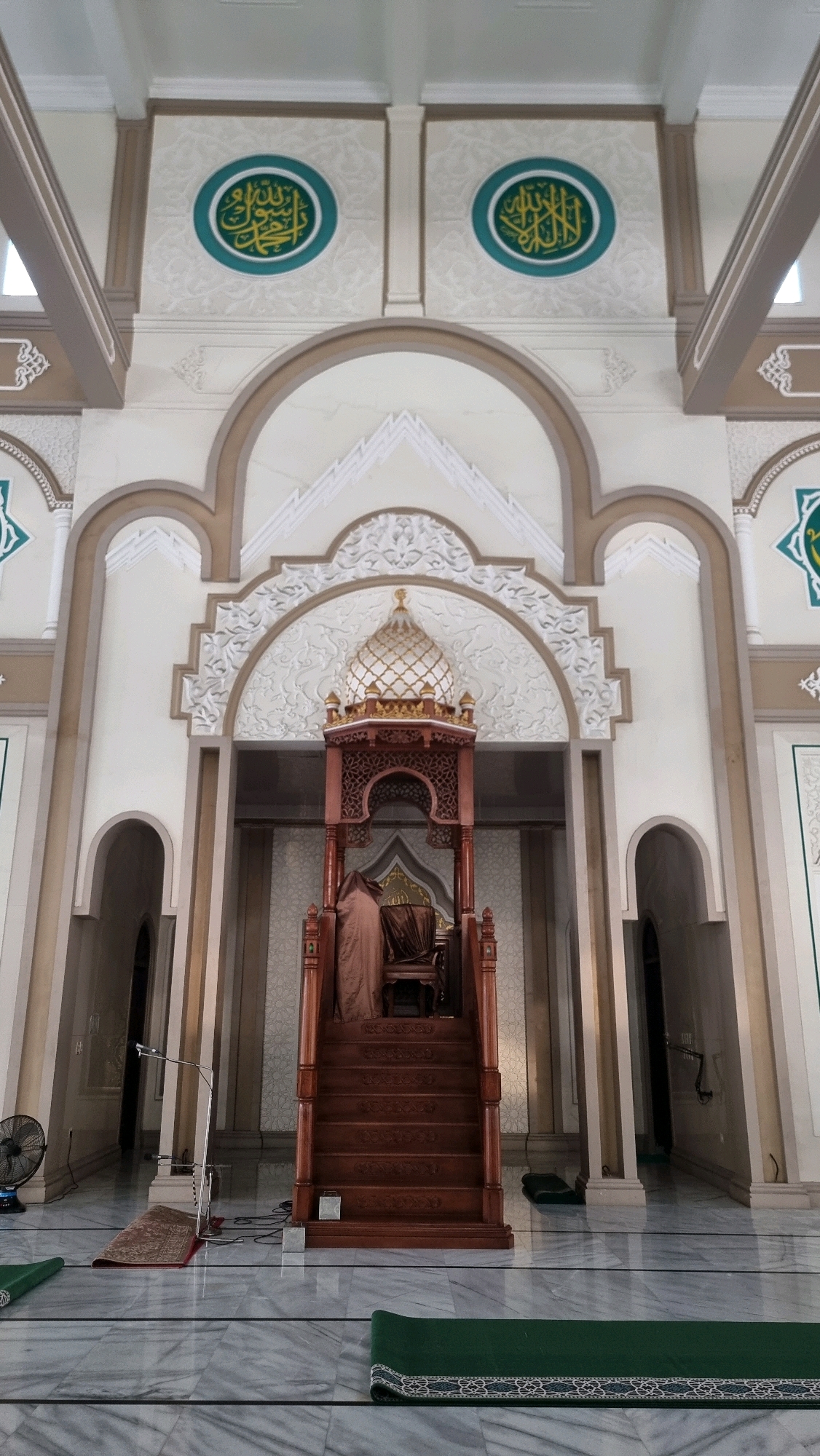 The image shows the interior of a mosque, likely in Lhokseumawe, Indonesia, with a prominent wooden minbar (pulpit) in the center. The minbar is elaborately carved and features a dome-like structure at the top. A brown cloth is draped over the seat of the minbar. The mosque's architecture includes arched doorways, decorative moldings, and circular green and gold Islamic calligraphy on the walls. The floor is made of polished marble with prayer mats laid out, including green ones in the foreground and a smaller red rug near a microphone stand and a fan on the left. The lighting is bright, illuminating the interior of the mosque.