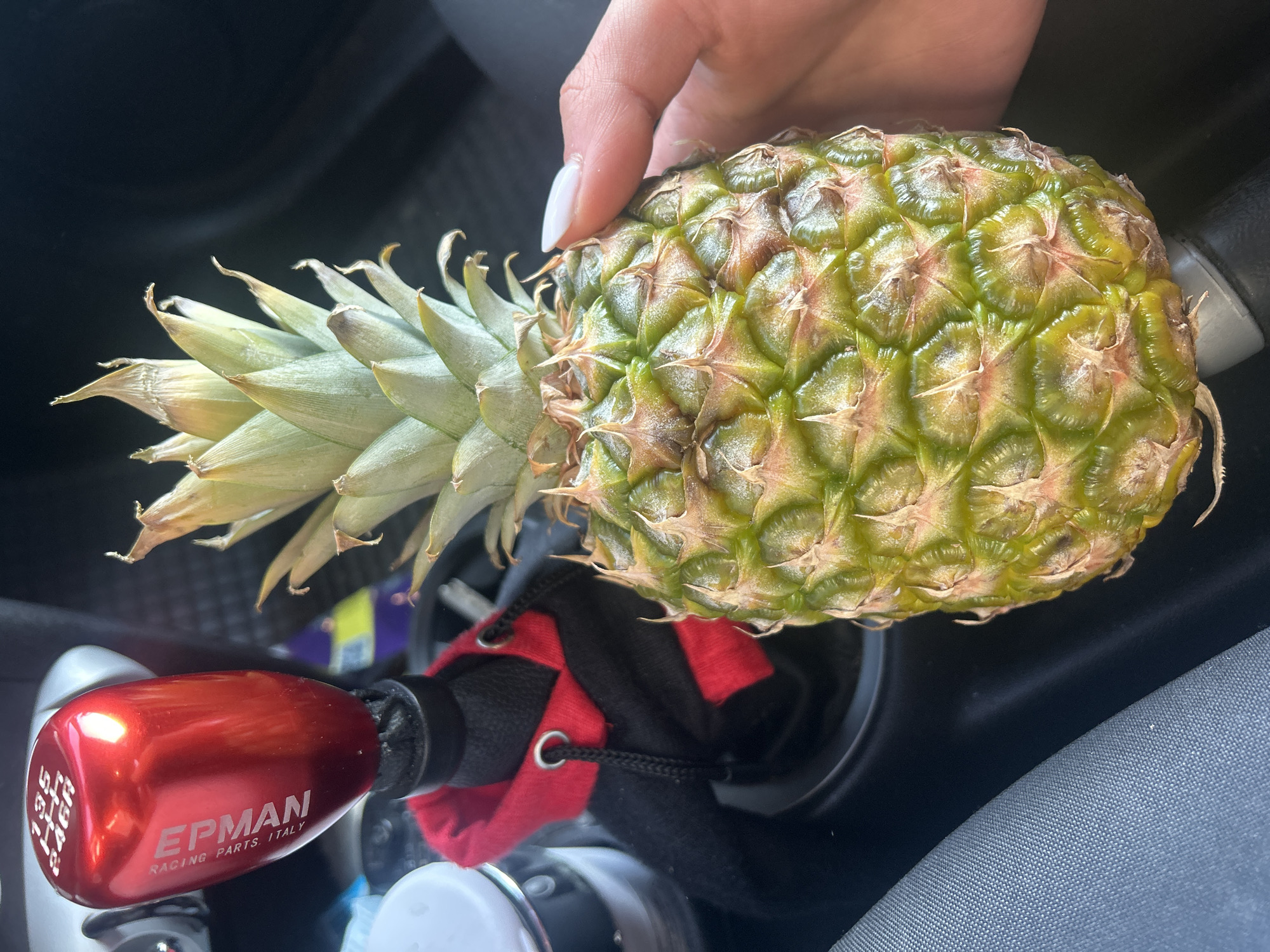 A hand holds a pineapple. The pineapple is positioned over a gear shift in a car. The gear shift is shiny red with "EPman Racing Parts Italy" on it. The surrounding environment of the car's interior includes a red and black sleeve around the base of the gear shift. The location in Burgas, Bulgaria cannot be determined from the image.
