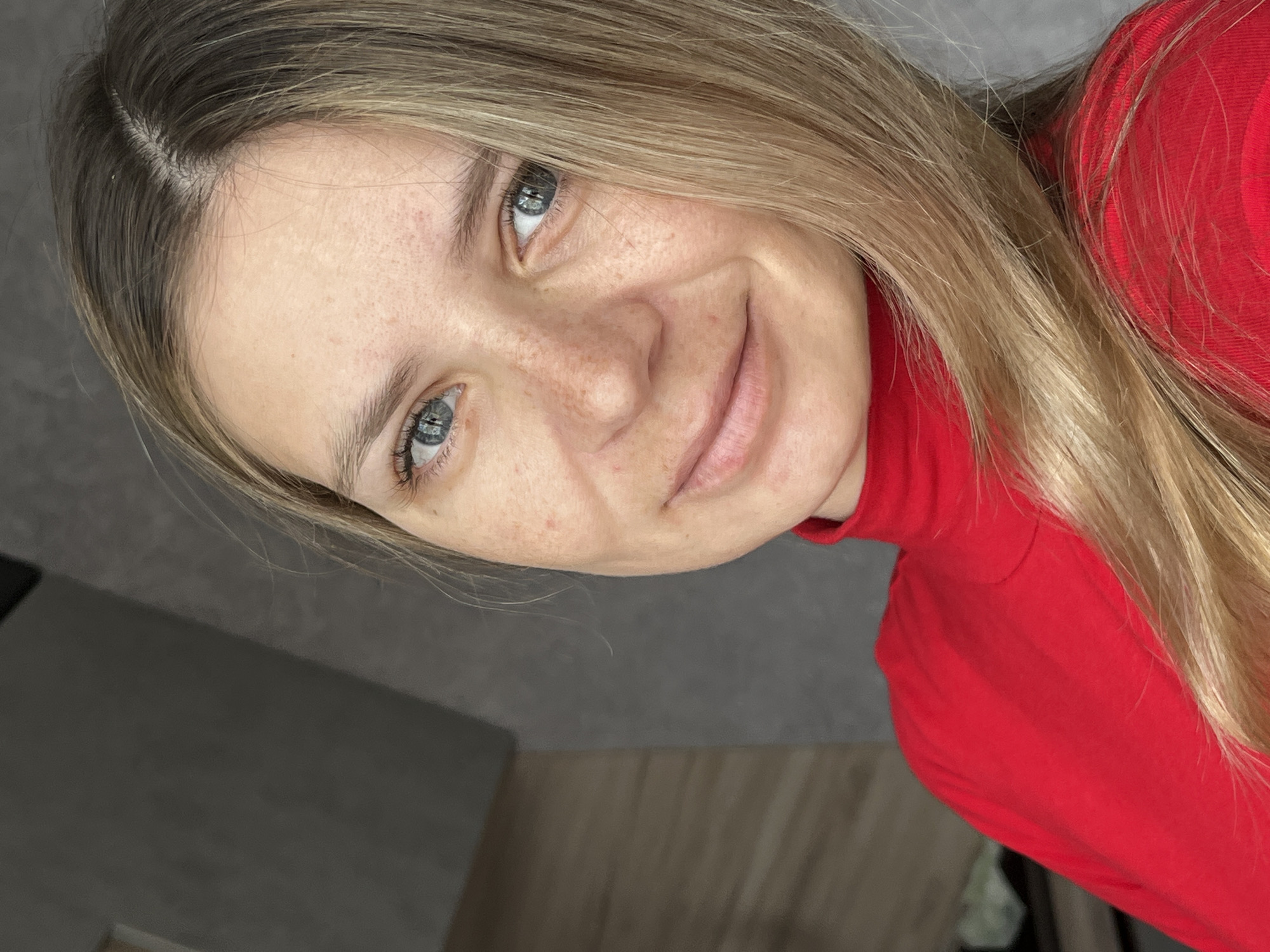 A close-up, tilted portrait shows a fair-skinned woman with freckles, light brown hair, and light blue eyes. She is wearing a red turtleneck sweater. The background is a mixture of gray and brown surfaces, possibly walls and furniture. No specific details indicate the location as Chaikovsky, Russia.