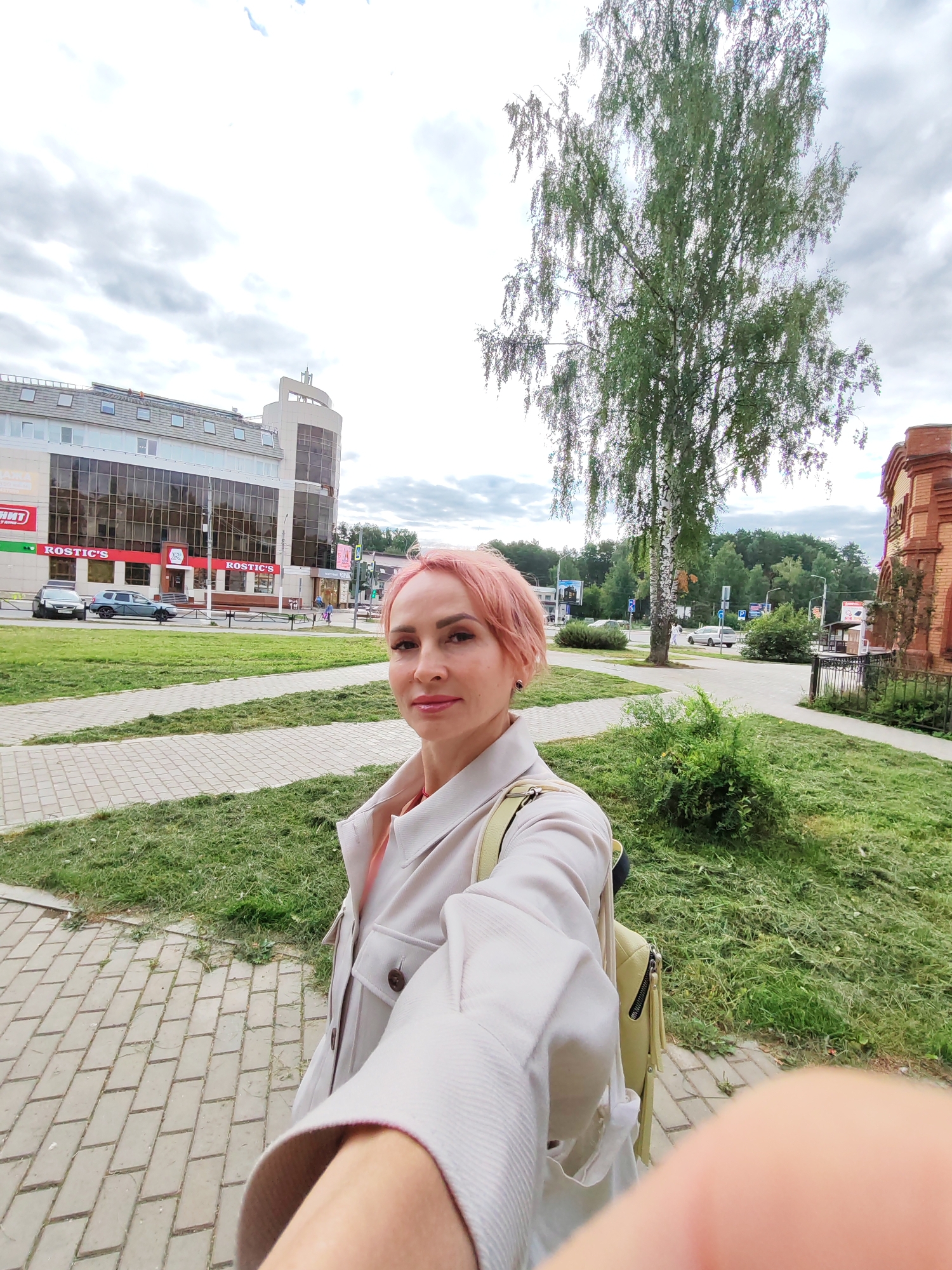 A woman with short pink hair takes a selfie outdoors in a city. She is wearing a cream-colored coat over an orange top and a light green backpack. The background shows a building with a glass facade and signs that read "ROSTIC'S." There are cars, a paved walkway, and a grassy area with a tall tree. The sky is cloudy. The location is Chaikovsky, Russia.