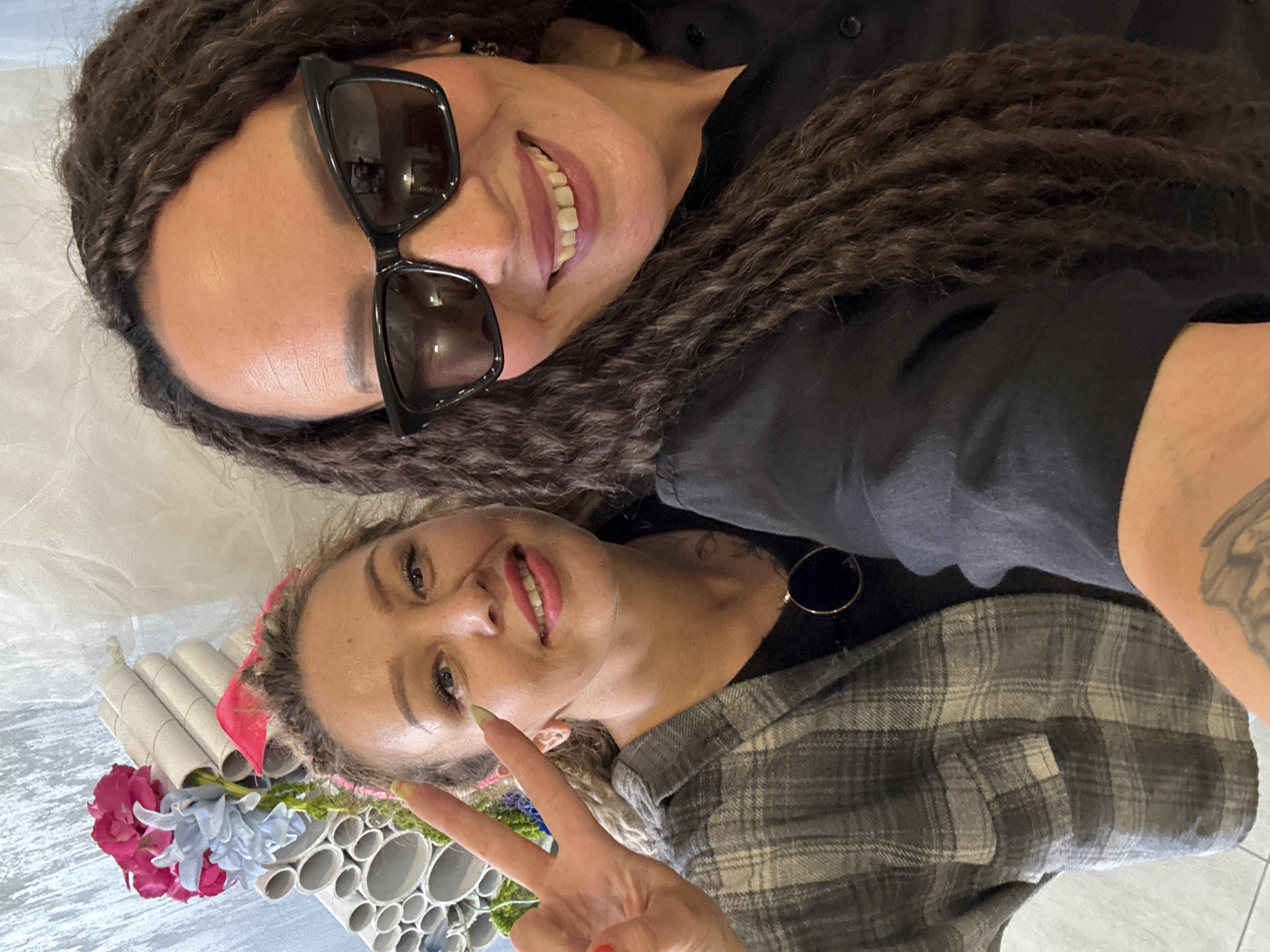 Two women are taking a selfie. The woman at the top of the image is wearing black sunglasses and a black top, and has long, dark, braided hair. The woman at the bottom is wearing a gray plaid shirt and has blonde hair in braids, with a red headband. She is holding up a peace sign with her right hand. There are flowers and what appear to be cardboard tubes in the bottom left corner. The location is Chaikovsky, Russia.