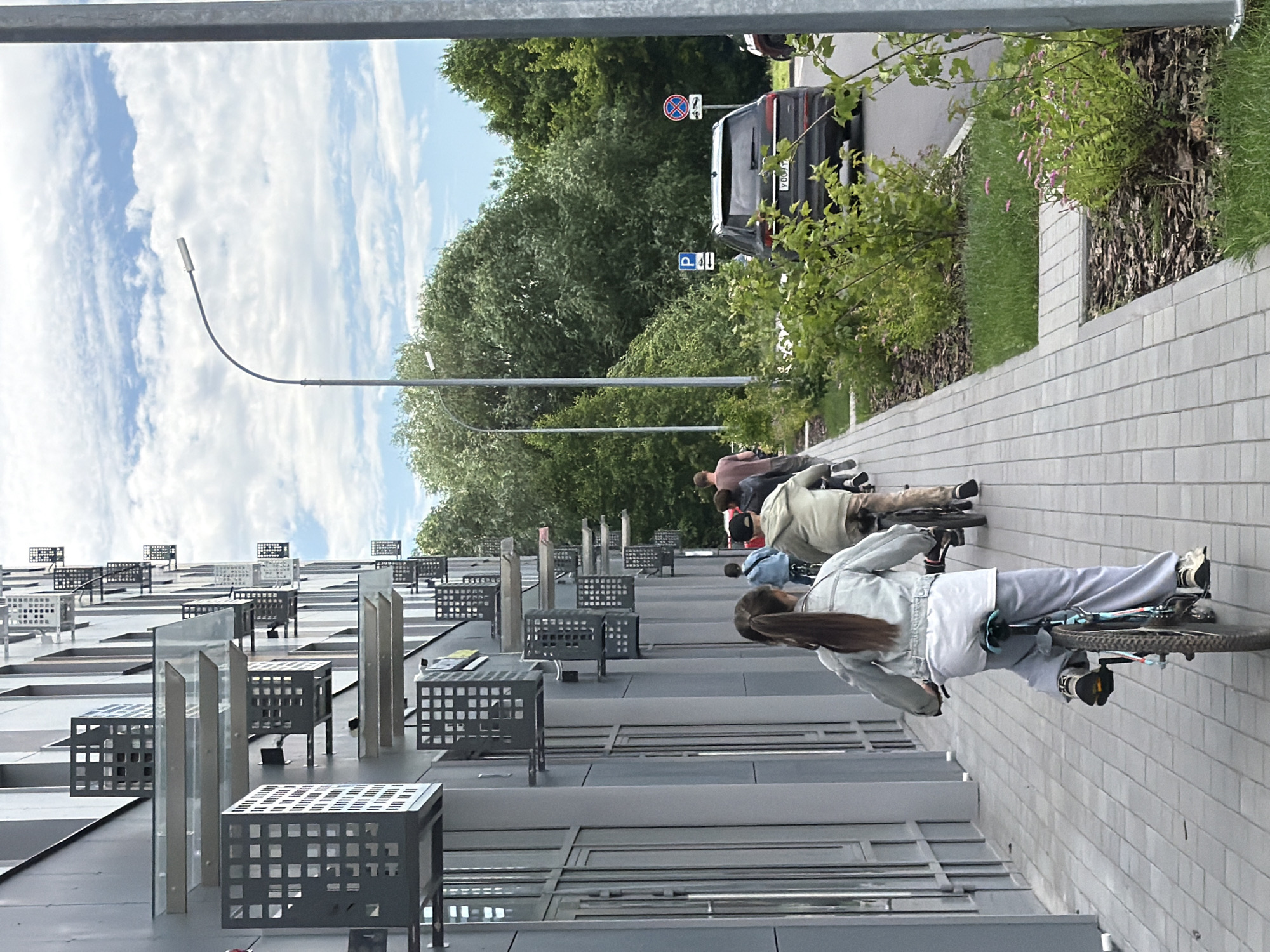 This media file captures an outdoor scene featuring several people on bicycles. The perspective is from the side, looking upwards towards the subjects and the sky. In the foreground, a woman on a bicycle is prominently visible, wearing a light blue denim jacket and grey pants. Her dark hair is long and reaches down her back. She is looking forward, and her bicycle is a dark color with black tires. Behind her, other individuals on bicycles are also visible, though less clearly. They appear to be riding in the same direction.
The setting includes a modern building with a grey facade and numerous square window-like structures or vents on its exterior. A sidewalk made of light-colored bricks runs alongside the building. Beyond the building and sidewalk, there are trees and greenery, suggesting an urban or suburban park-like environment. A black SUV is parked on a road in the background, with a traffic sign visible above it. The sky is blue with scattered white clouds.
The image appears to be taken during the daytime, with good lighting. The overall mood seems relaxed and casual, consistent with people enjoying a bike ride. There is no visible text in the image that provides specific location information, though the style of the building and the presence of a street with a car suggest a developed area.