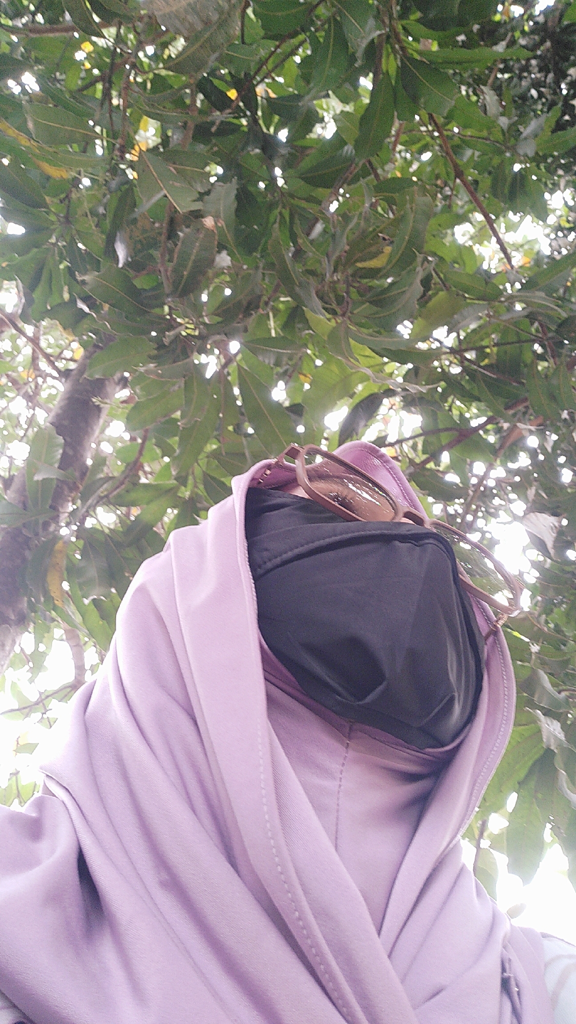 A low-angle, medium shot captures a person from the chest up, looking upwards through a canopy of green leaves. The person is wearing a light purple hijab and a black face mask, with a pair of brown-rimmed glasses resting on top of the hijab. The background consists of densely packed, dark green leaves and branches, with bright, diffused sunlight filtering through, suggesting an outdoor setting, possibly in Karya Sakti, Indonesia, under a tree. The focus is on the person's attire and the natural environment.