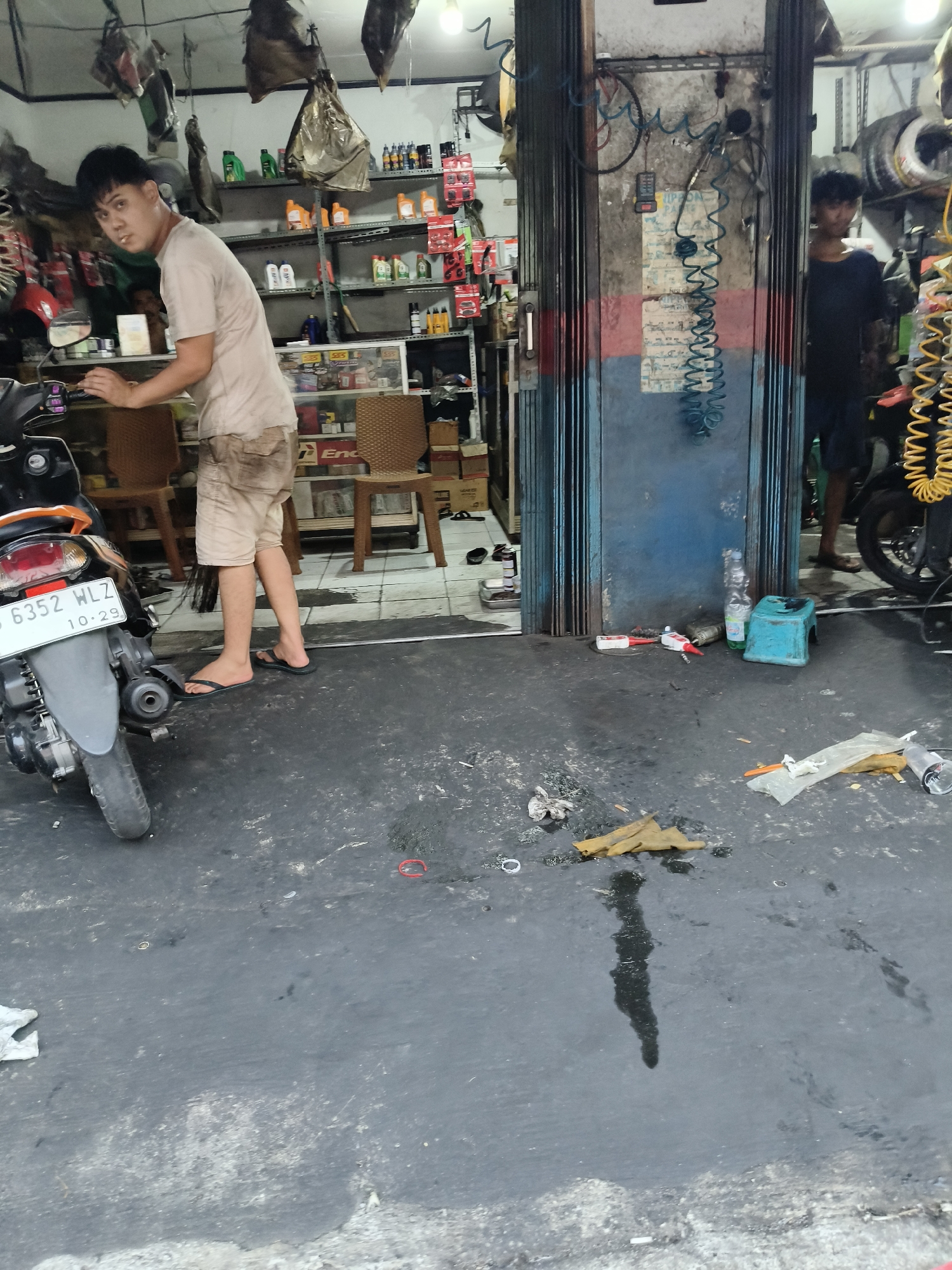 A young male in tan shorts and a light-colored t-shirt stands in a cluttered motorcycle repair shop. The shop has shelves filled with various automotive products, including oil bottles and car parts. A motorcycle is parked in the foreground. A second male, wearing a dark shirt and shorts, is visible in the background. The floor of the shop is dark and appears to be wet or oily, with various tools and debris scattered around. The scene suggests a working environment in a busy part of South Tangerang, Indonesia.