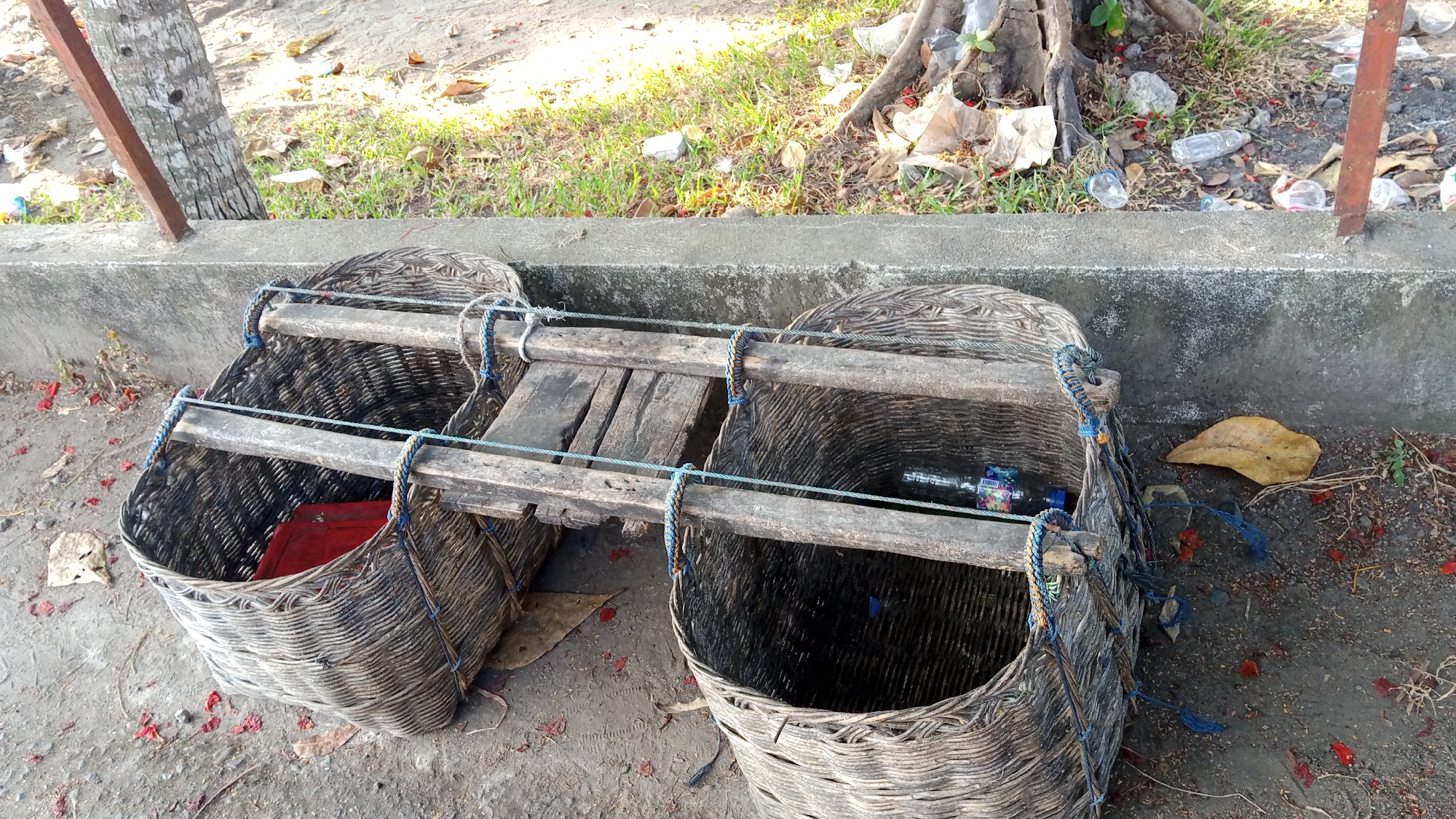 The image depicts two large, woven baskets, possibly used for carrying goods, positioned side-by-side outdoors. They are made of dark, natural fibers and are reinforced with wooden planks across their tops, secured by blue and yellow braided ropes. The basket on the left contains a red object, while the basket on the right holds a plastic bottle and other small items. Both baskets rest on a gray, concrete surface. Behind the baskets, a textured concrete wall runs horizontally, and beyond that, a grassy area with scattered leaves, small red flowers, and some discarded plastic bottles is visible. A tree trunk with exposed roots is present in the background, along with a weathered wooden post. The lighting suggests a sunny day. The scene is located in Julikeude Dua, Indonesia, as indicated by the prompt.
