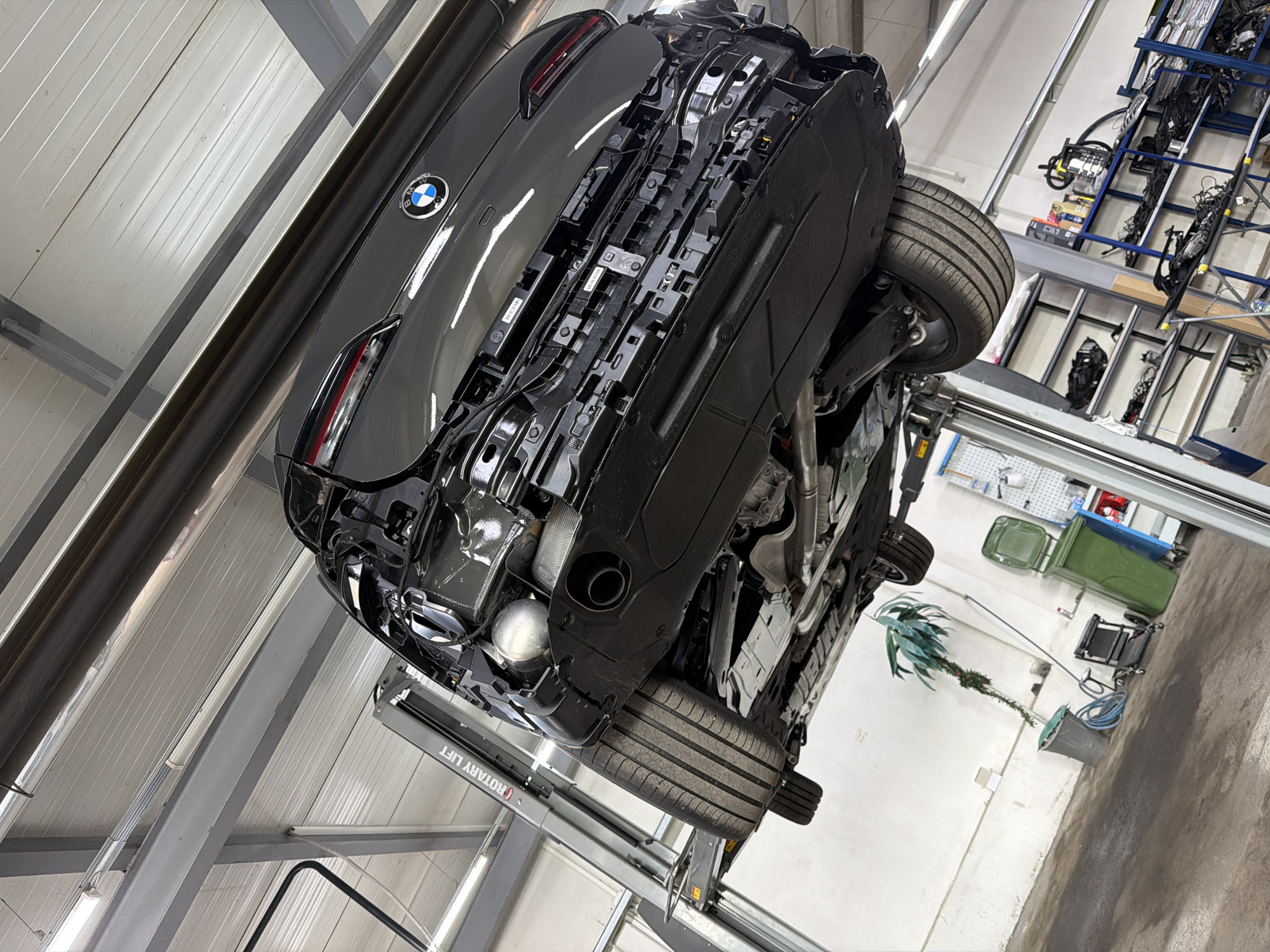 A low-angle photograph, rotated approximately 45 degrees counter-clockwise, captures the underside and rear of a dark grey BMW automobile elevated on a hydraulic car lift within an indoor automotive workshop or garage. The car, prominently featuring its circular BMW emblem on the trunk and a distinctive taillight design, is positioned diagonally across the frame. Its rear bumper assembly appears partially disassembled or exposed, revealing internal components beneath the bodywork.
The car's rear wheels, fitted with tires, are clearly visible, with the suspension and exhaust system components exposed beneath the vehicle. The grey car lift, identified by the text "ROTARY LIFT" (visible reversed due to the image orientation) on its mechanism, supports the vehicle.
The setting is a well-lit garage, with white corrugated metal walls and ceiling panels, and grey support beams. Fluorescent light tubes illuminate the space. In the background, various workshop items are discernible: a multi-tiered shelving unit laden with wires and possibly other automotive parts, green industrial trash bins, a wheeled cart, a long-stemmed plant in a pot, and hoses coiled on the floor. A pegboard with tools can also be seen further back. The overall impression is an active service bay where the car is undergoing maintenance or repair in Burgas, Bulgaria. No people are visible.