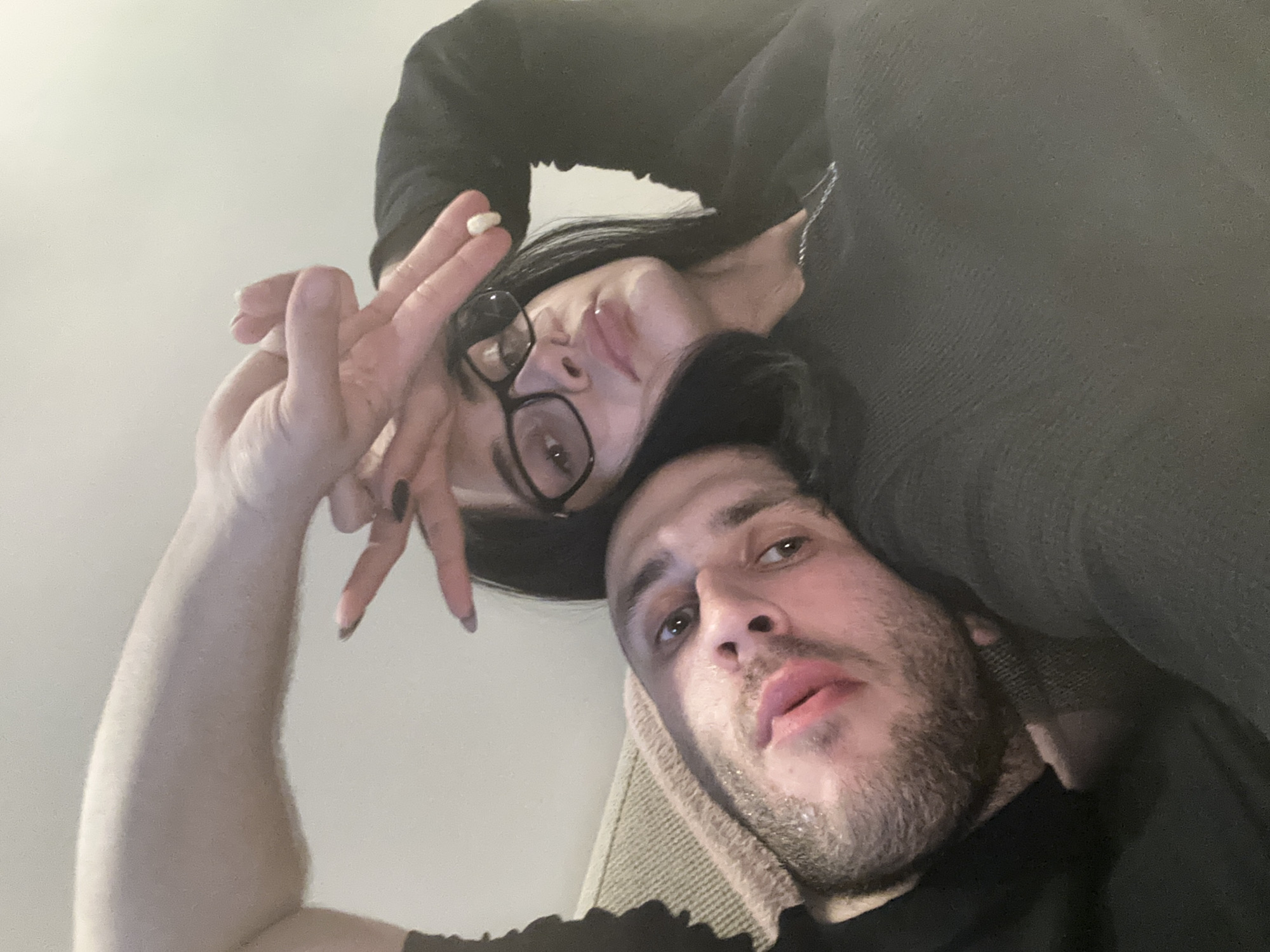 Two people are pictured in a selfie-style photograph, appearing to be lying down. The man has a beard and dark hair, and is wearing a dark shirt. The woman is wearing glasses, has dark hair, and is holding a small white object between her fingers. The setting is likely indoors. The location in Burgas, Bulgaria, cannot be determined based solely on the visual information provided in the image.