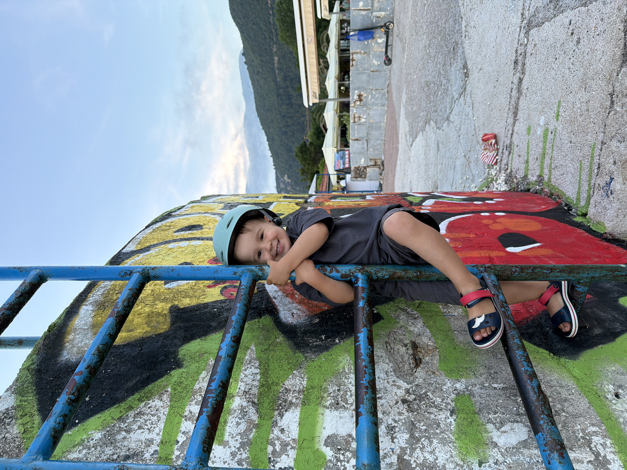 The image displays a joyful young child lying horizontally on a weathered blue metal structure, positioned against a concrete wall adorned with vibrant graffiti. The child, appearing to be a toddler or preschooler, wears a light blue helmet, a dark grey t-shirt, and dark shorts. They are smiling broadly at the camera and giving a thumbs-up gesture. Their feet, wearing navy blue sandals with red straps and white shark motifs, are visible through the metal rungs.
The concrete wall behind the child features bold, colorful graffiti in shades of red, black, yellow, and green, showing signs of peeling paint. In the background, under a partly cloudy, bright sky, a range of green, tree-covered mountains dominates the horizon. Below the mountains, several buildings with light-colored awnings and parked scooters are visible, suggesting a commercial or residential area along a coastline. The soft lighting indicates it is likely late afternoon or early evening. The scene captures a playful moment, with the child expressing clear happiness. The location context of Budva, Montenegro, aligns with the coastal mountain scenery. No discernible text is legible within the image.