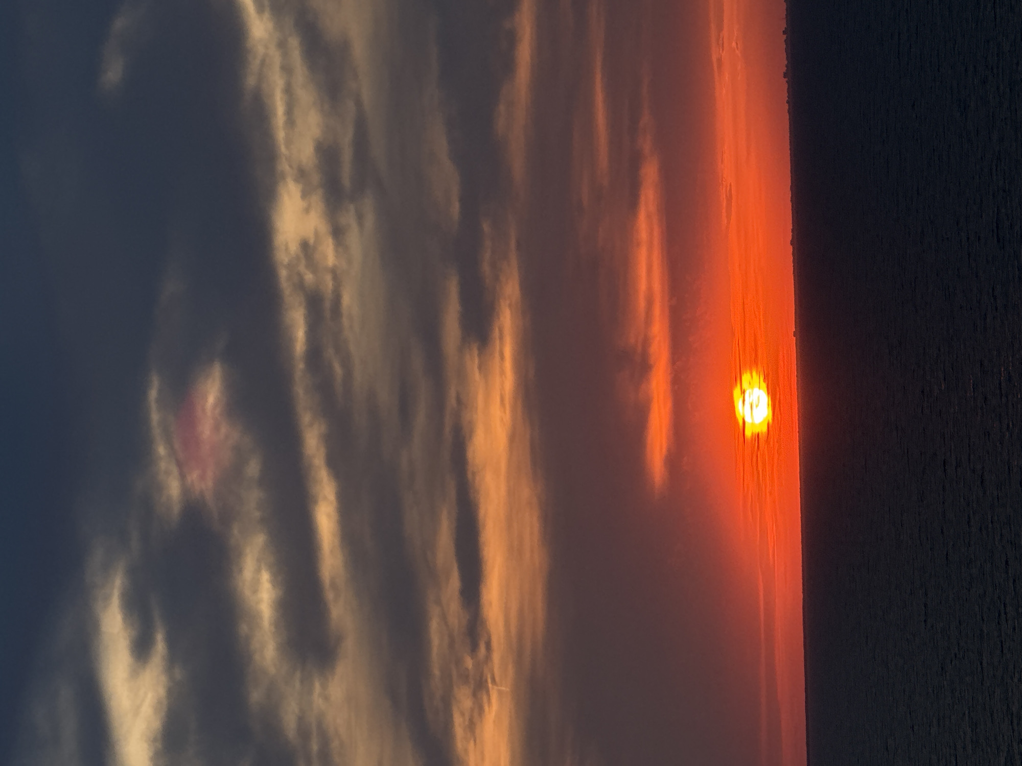 The image depicts a vibrant sunset over a body of water, possibly a sea or ocean, with a dark horizon line suggesting land in the distance. The sky is a dramatic display of colors, transitioning from deep blues and grays at the top to fiery oranges and reds closer to the horizon. Clouds are present, catching the sunlight and adding texture to the sky. A bright, circular sun is visible, partially obscured by clouds, casting a warm glow. The water below is dark and calm, reflecting the colors of the sky. The overall impression is one of natural beauty and tranquility. There are no people or specific landmarks identifiable in the image that would allow for pinpointing a location within Unknown, Italy.