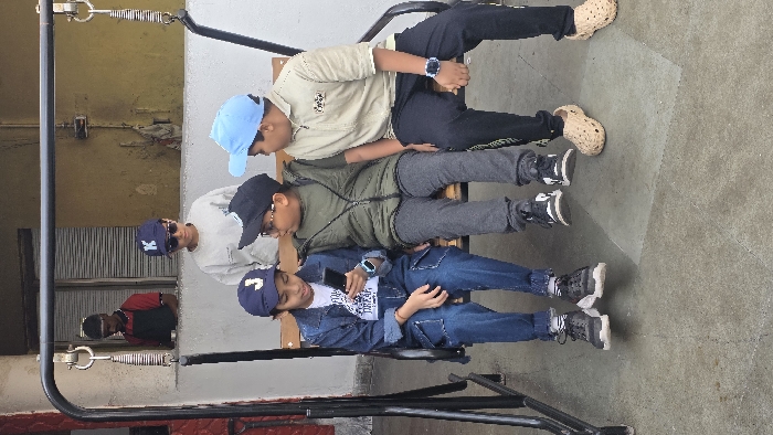 Four boys sit on a swing. The boy on the top wears a light tan shirt and dark pants. The second boy wears a green jacket and gray pants. The third wears a light-colored shirt and dark sunglasses. The bottom boy wears a denim jacket and jeans, holding a phone. Each boy wears a different colored hat, ranging from blue to black. A man in a red shirt stands in a window in the background.